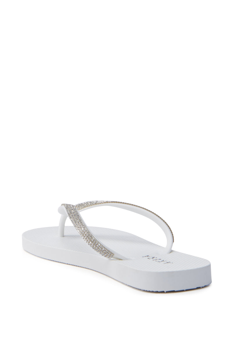 The AKIRA LORETA WHITE THONG FLIP FLOP features a white thong design with sparkling rhinestone-adorned straps, displayed at an angle on a plain background. It has a slightly textured PVC sole for grip.