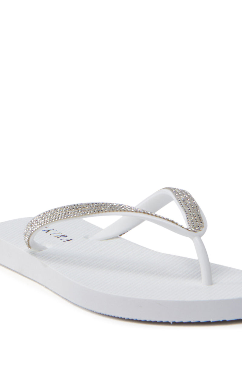 A close-up of the AKIRA LORETA WHITE THONG FLIP FLOP showcases its simple yet elegant design, featuring gleaming rhinestone embellishments on PVC straps. It lies on a plain white background, highlighting its charm.