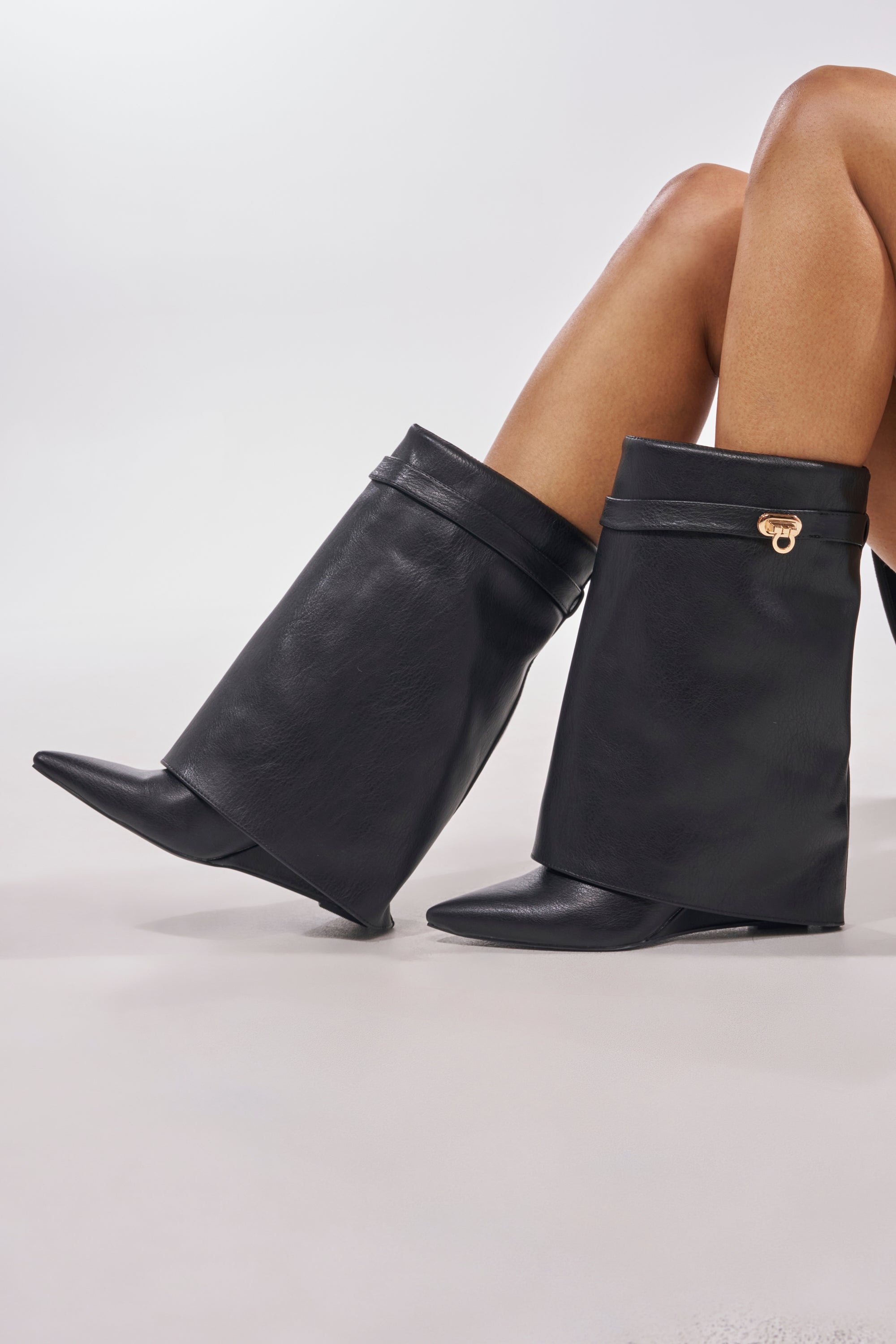 A person with crossed legs wears the AZALEA WANG LOUISA BLACK PU WEDGE BOOTIE, featuring eye-catching black faux leather, wide flared shafts, pointed toes, and a small gold buckle. The background is a plain light color. 0