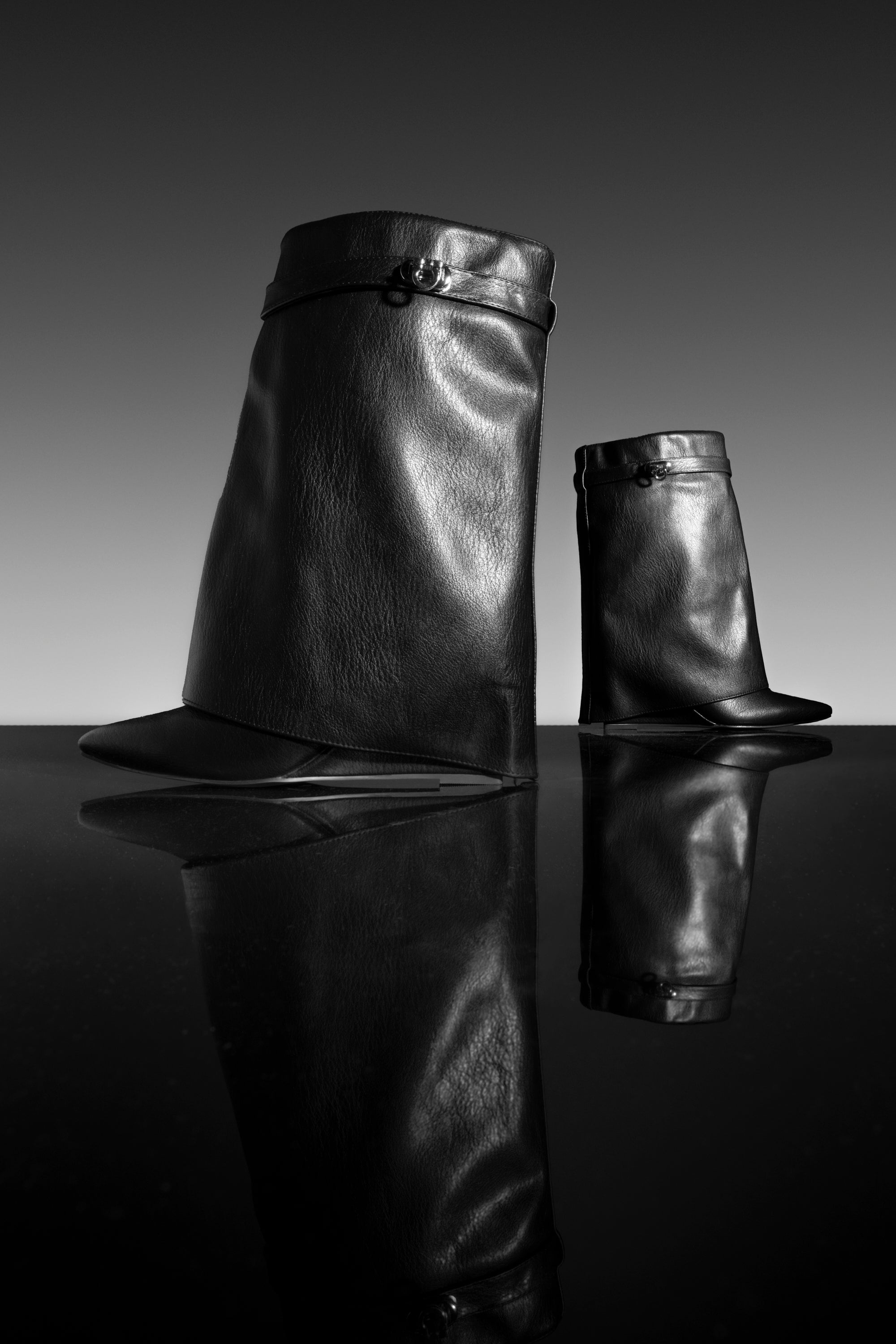 Two AZALEA WANG LOUISA BLACK PU WEDGE BOOTIES, crafted from sleek faux leather and featuring an abstract, sculptural silhouette, are displayed on a glossy surface that mirrors their forms. The background transitions from dark to light, enhancing the dramatic contrast of the scene.