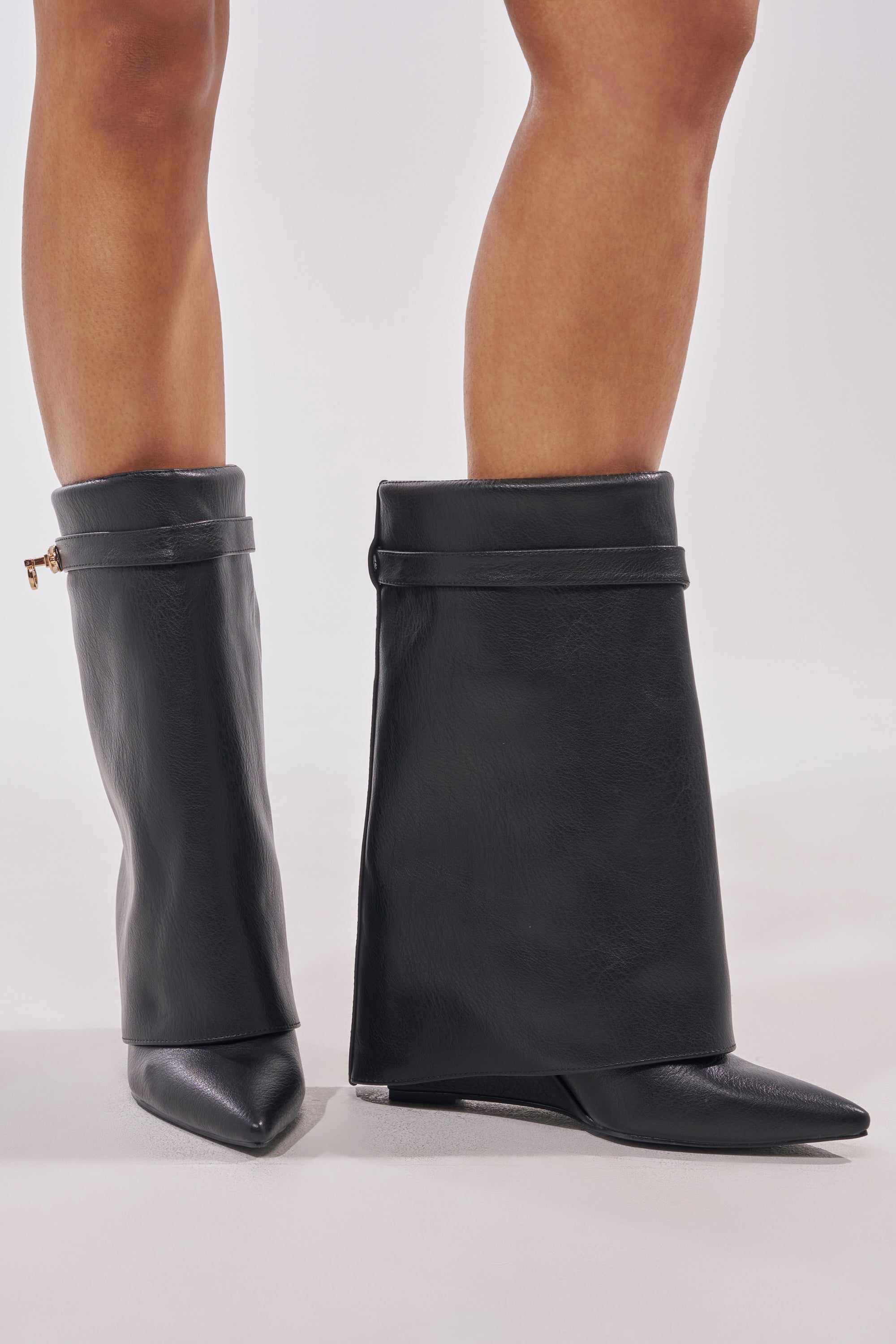 A person wears the AZALEA WANG LOUISA BLACK PU WEDGE BOOTIE, featuring wide, flared tops, pointed toes, and a bold silhouette. The striking black faux leather booties showcase a modern, avant-garde design on a light-colored surface. 1