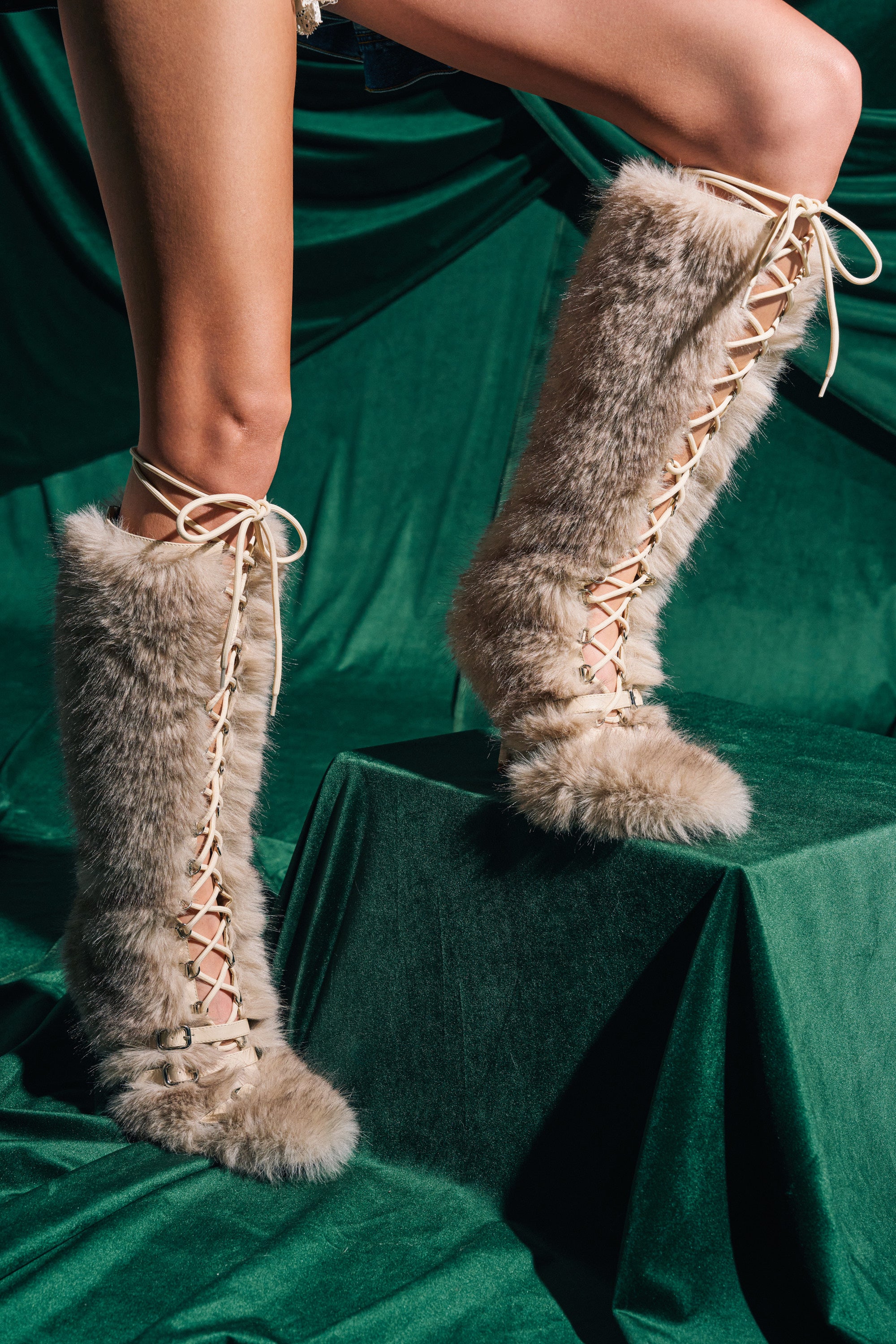 Only the legs are visible of a person standing on green velvet, with matching green draped fabric behind, wearing the AZALEA WANG LOURDANNE IVORY FUR LACE UP BOOT—knee-high faux fur boots with stiletto heels.