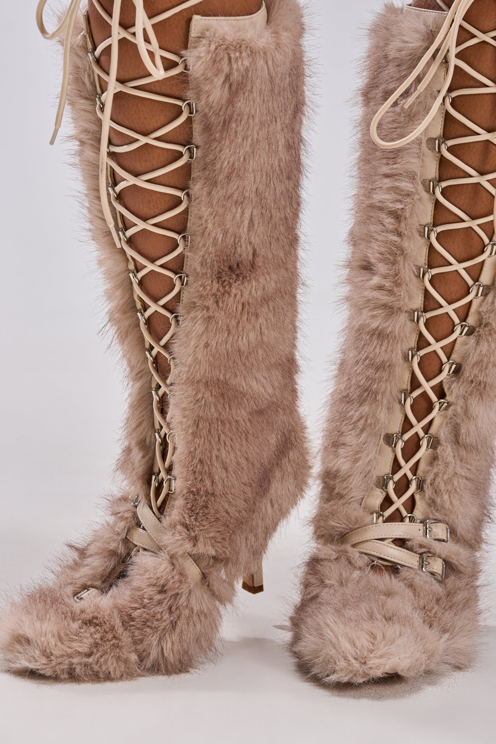Close-up of the AZALEA WANG LOURDANNE IVORY FUR LACE UP BOOT—knee-high with beige laces, buckles, pointed toes, and thin stiletto heels. Covered in light ivory faux fur for a chic and cozy winter style.