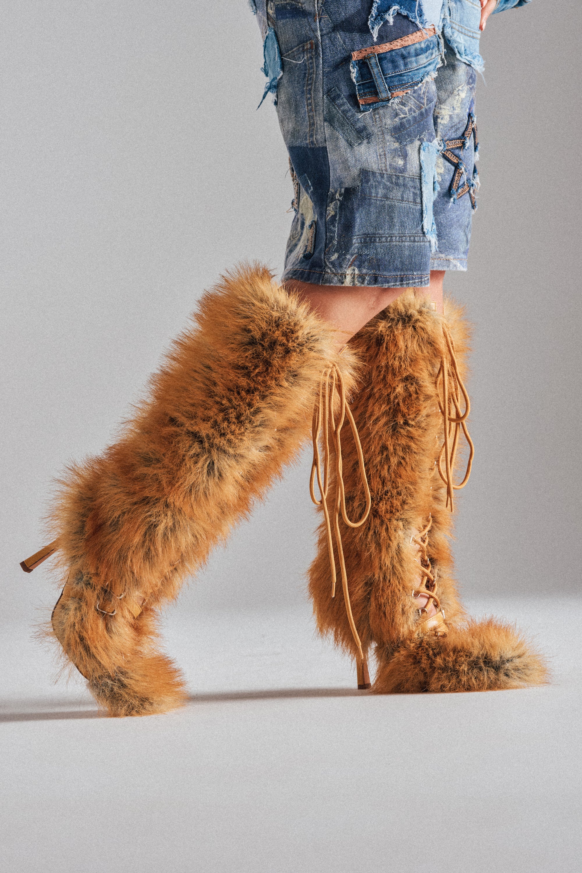 Wearing the AZALEA WANG LOURDANNE Tan Fur Lace Up Stiletto Boot with distressed denim shorts and a patchwork denim jacket, a person stands against a neutral background.