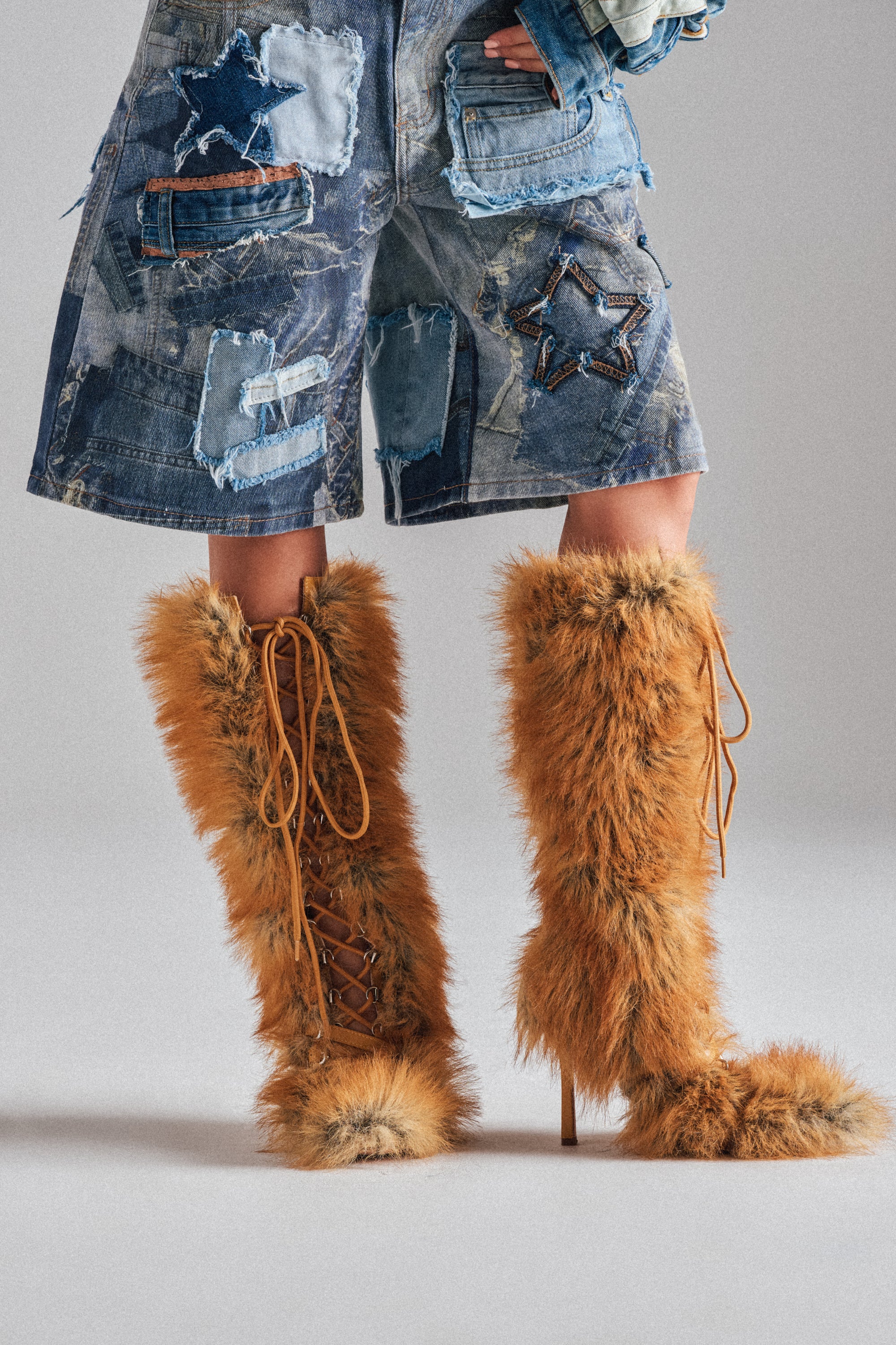 A person in knee-length patchwork star denim shorts and knee-high AZALEA WANG LOURDANNE TAN FUR LACE UP STILETTO BOOT stands on a plain light background.