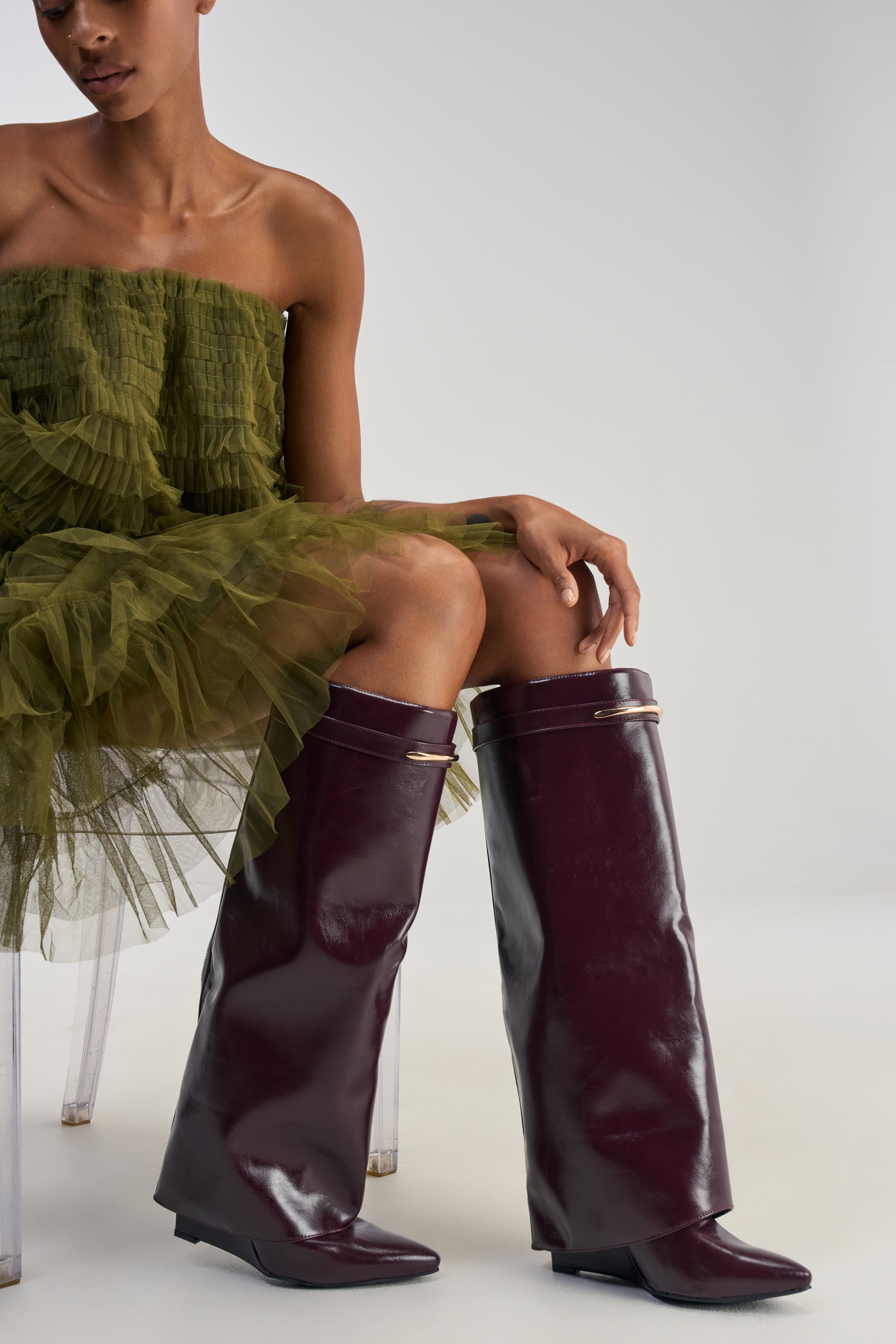 A person wearing a green tulle dress sits on a clear chair, showcasing the AZALEA WANG LOVE BURN BURGUNDY SHARK WEDGE BOOT—shiny, knee-high footwear featuring pointed toes and gold accents.
