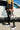 A person wearing a yellow crop top, black puffy mini skirt, and AZALEA WANG OH MY LOVE Faux Leather Thigh High Flatform Sneaker Boots with 4-Way Stretch in Black is holding a white and black handbag with the number 14 on it. They stand in front of a white car.