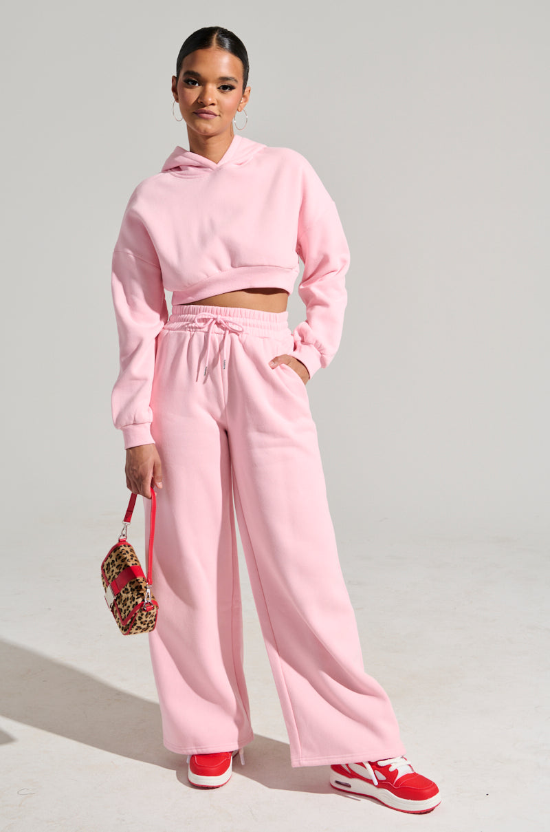 Wearing the LOVER WIDE LEG SWEATPANT in a pink color, a person stands confidently with a matching cropped hoodie. The high-rise fit and elastic waistband of the sweatpants are complemented by a small leopard print handbag they hold, while red and white sneakers complete their look against a plain background. 0