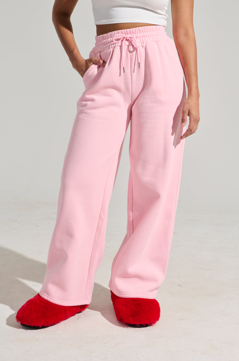 A person wearing a white crop top, LOVER WIDE LEG SWEATPANT in pink with a high-rise fit and drawstring waist, and furry red slippers stands with one hand in their pocket against a plain background. The photo captures a casual, laid-back outfit style. 1