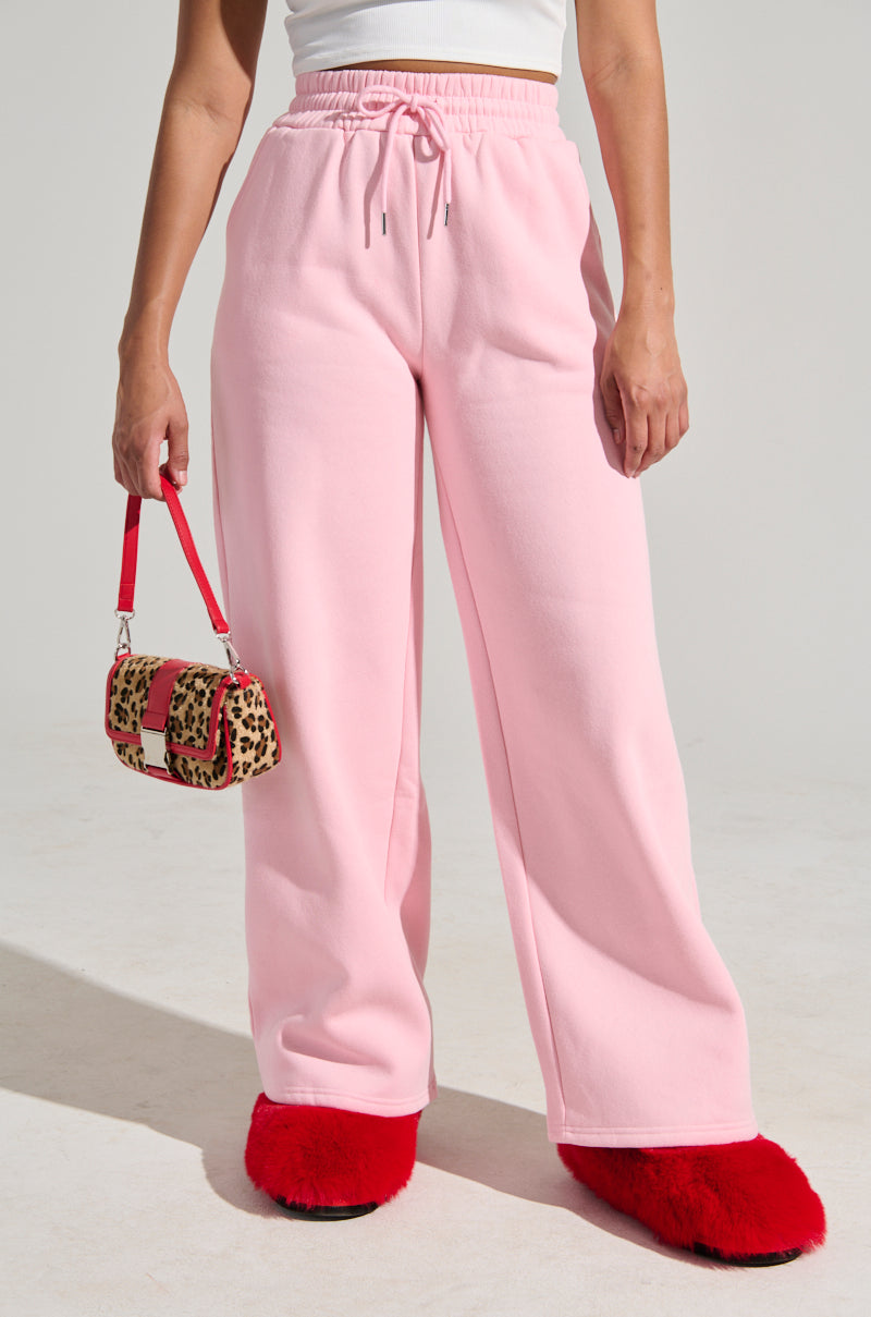 A person in light pink LOVER WIDE LEG SWEATPANT and a white top holds a small leopard print handbag with a red strap. They wear bright red fuzzy slippers. The background is a plain light color. 2