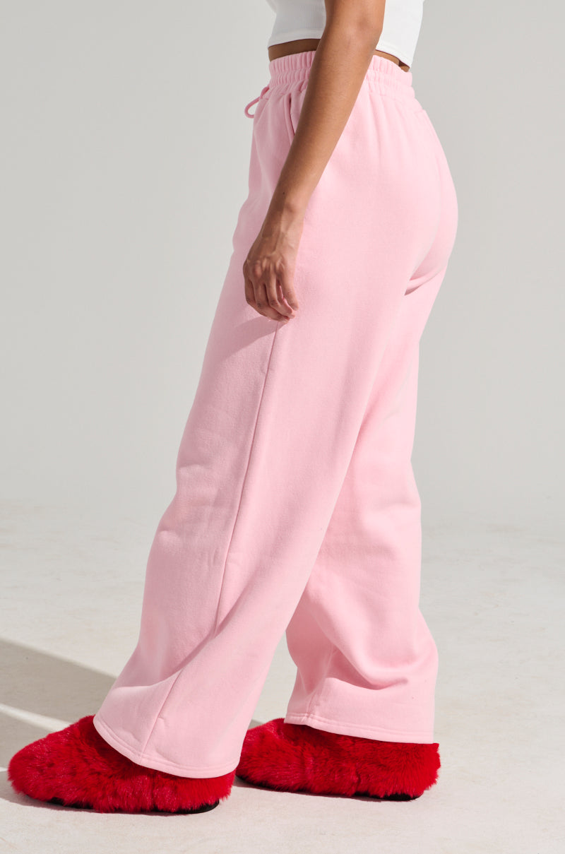 A person dressed in the LOVER WIDE LEG SWEATPANT in pink, featuring an elastic waistband, and a white top is standing sideways. They are wearing red, fluffy slippers on their feet against a plain, light-colored background. 3