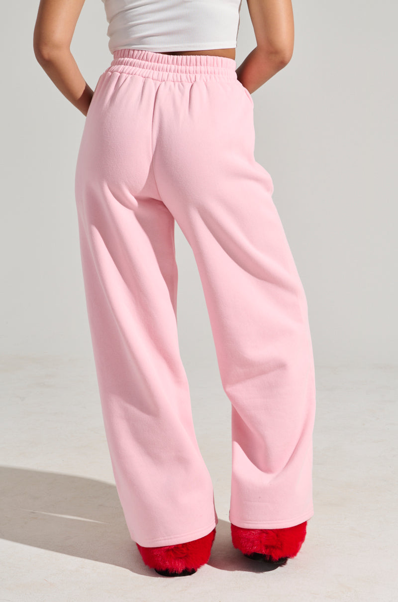 A person wearing LOVER WIDE LEG SWEATPANT in light pink with an elastic waistband and a white top along with red fluffy slippers stands facing away. The scene is set against a plain white background. 4