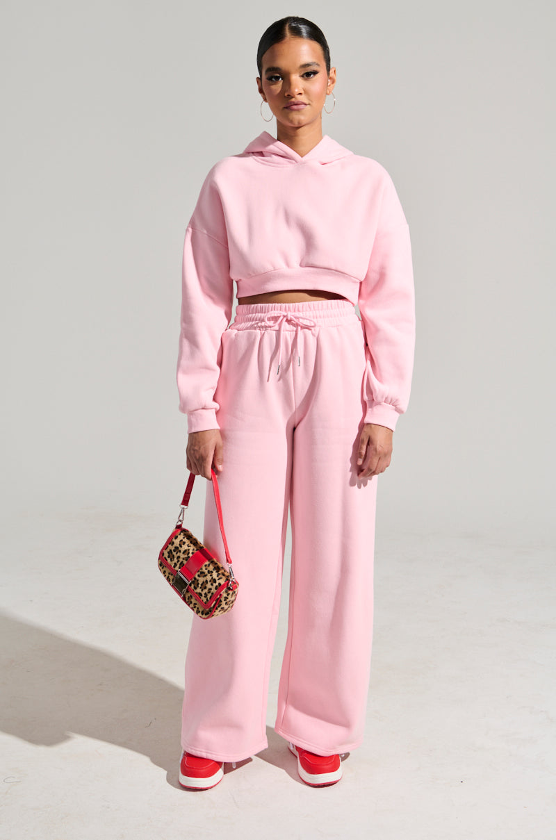 A person stands wearing a pink high-rise fit hoodie paired with the LOVER Wide Leg Sweatpant featuring an elastic waistband. They hold a small leopard print handbag and sport red sneakers. The background is plain and well-lit. 5