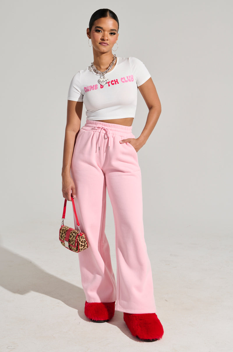 A woman poses confidently in a white cropped T-shirt and LOVER WIDE LEG SWEATPANTS featuring a high waist with an elastic waistband. She holds a small animal print handbag and sports bold red furry slippers. Her hair is tied back, accessorized with necklaces, completing her stylish high-rise fit. 6