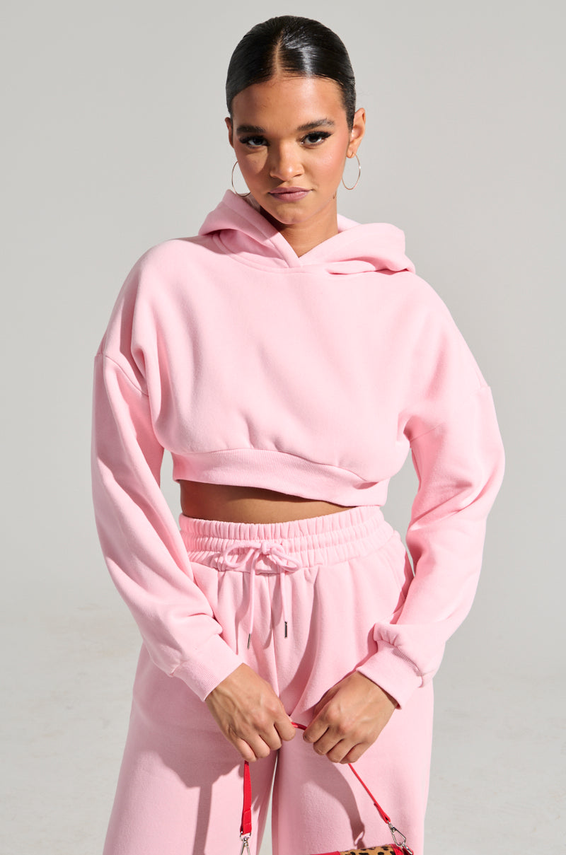 A person with dark hair tied back is posing confidently in a LOVER CROPPED HOODIE paired with matching sweatpants. They hold a small strap, exuding style against a plain, light-colored background that enhances the monochrome finish of their cozy ensemble.
