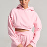 A person with dark hair tied back is posing confidently in a LOVER CROPPED HOODIE paired with matching sweatpants. They hold a small strap, exuding style against a plain, light-colored background that enhances the monochrome finish of their cozy ensemble.