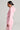 A person in profile is dressed in a pink LOVER CROPPED HOODIE tracksuit. Their hair is neatly tied back, and they wear hoop earrings. The neutral background accentuates the soft color and casual style of the pastel outfit.