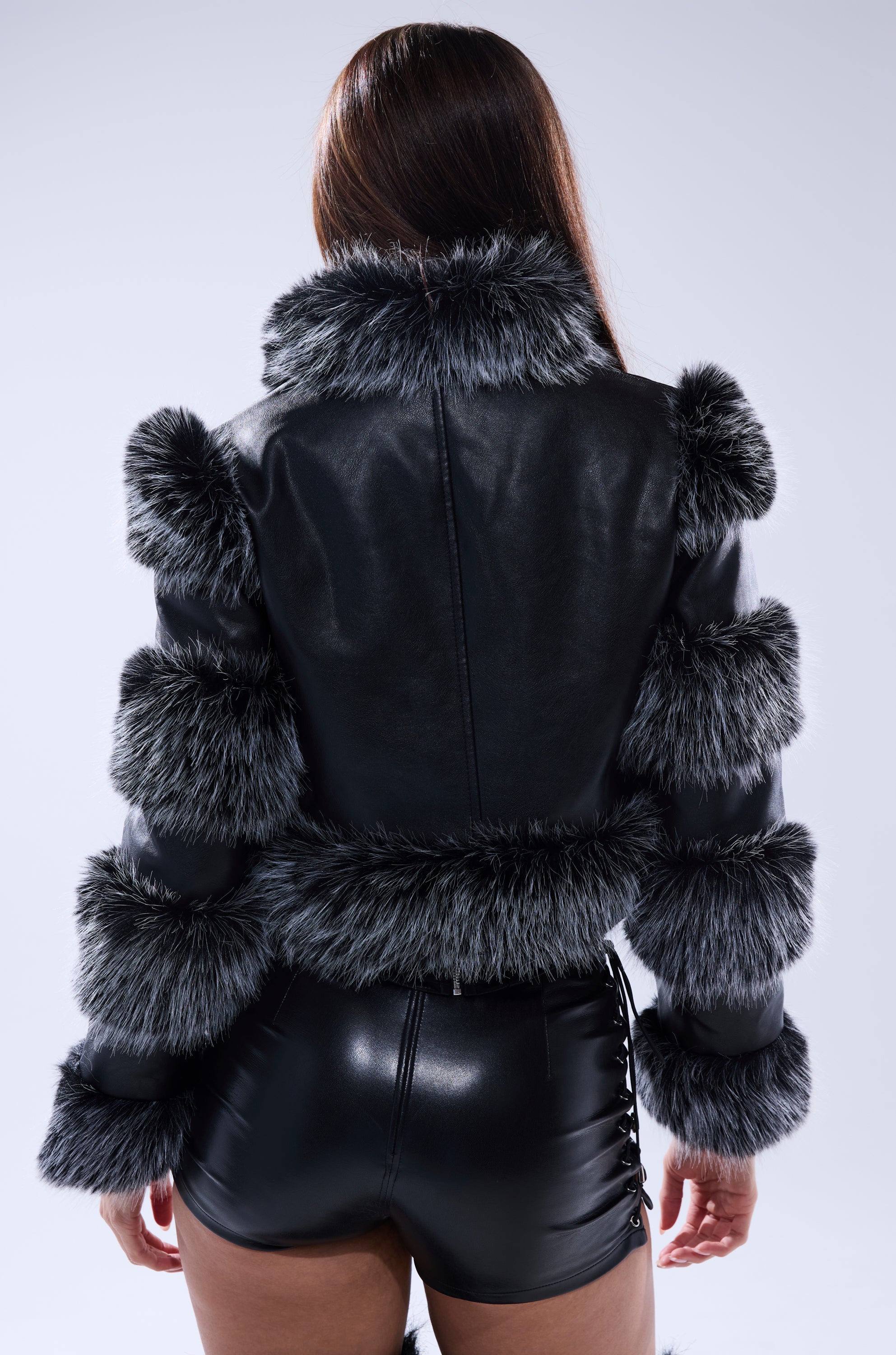 A person with long brown hair, seen from behind, wears the LOVERBOY FAUX FUR MOTO JACKET IN BLACK with matching faux leather shorts. The background is plain and light-colored.