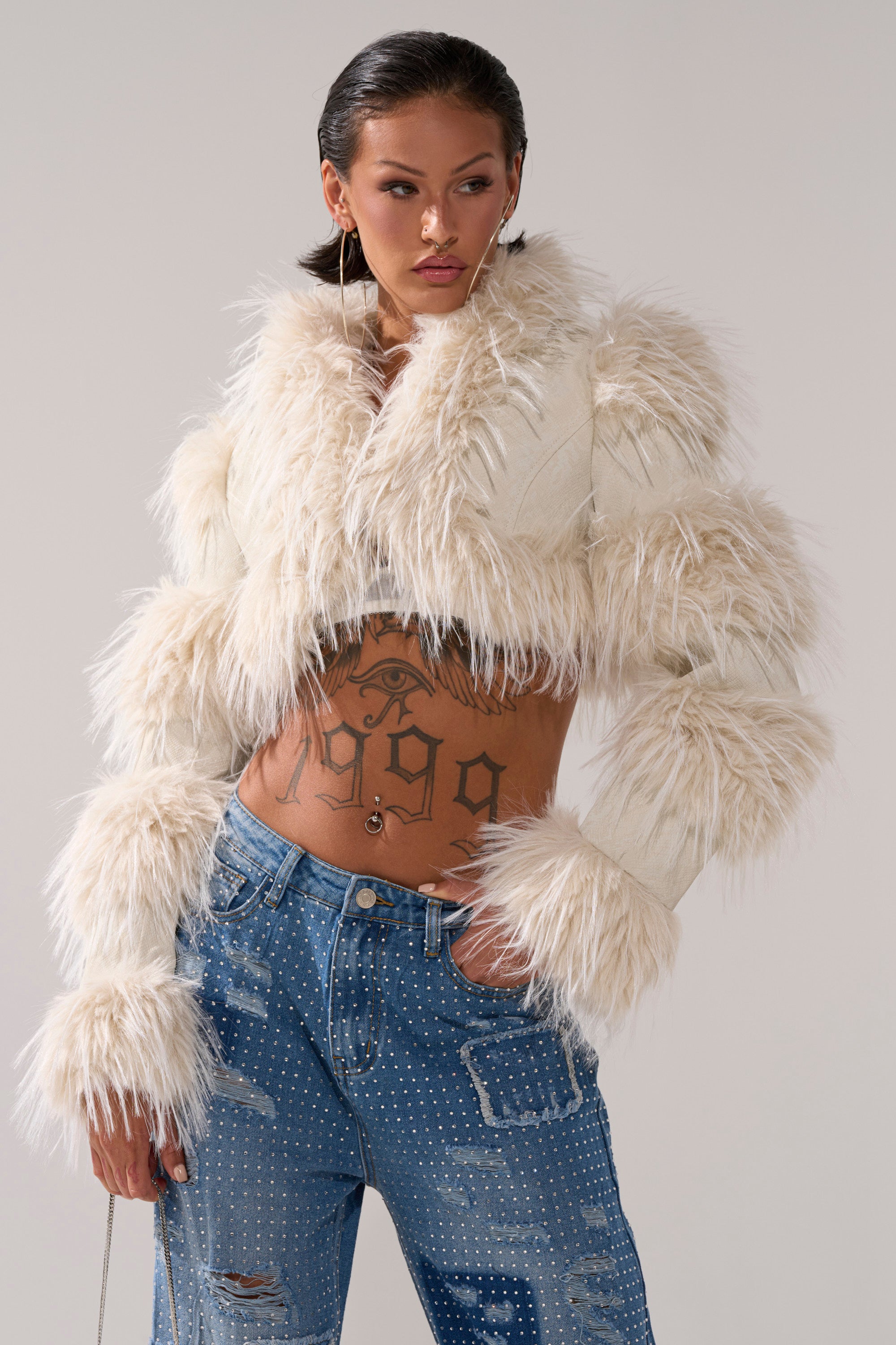 A woman wears the LOVERGIRL ULTRA CROP FUR TRIM MOTO IN IVORY with blue rhinestone jeans, styled with wet-look hair, hoop earrings, and a bold stomach tattoo.