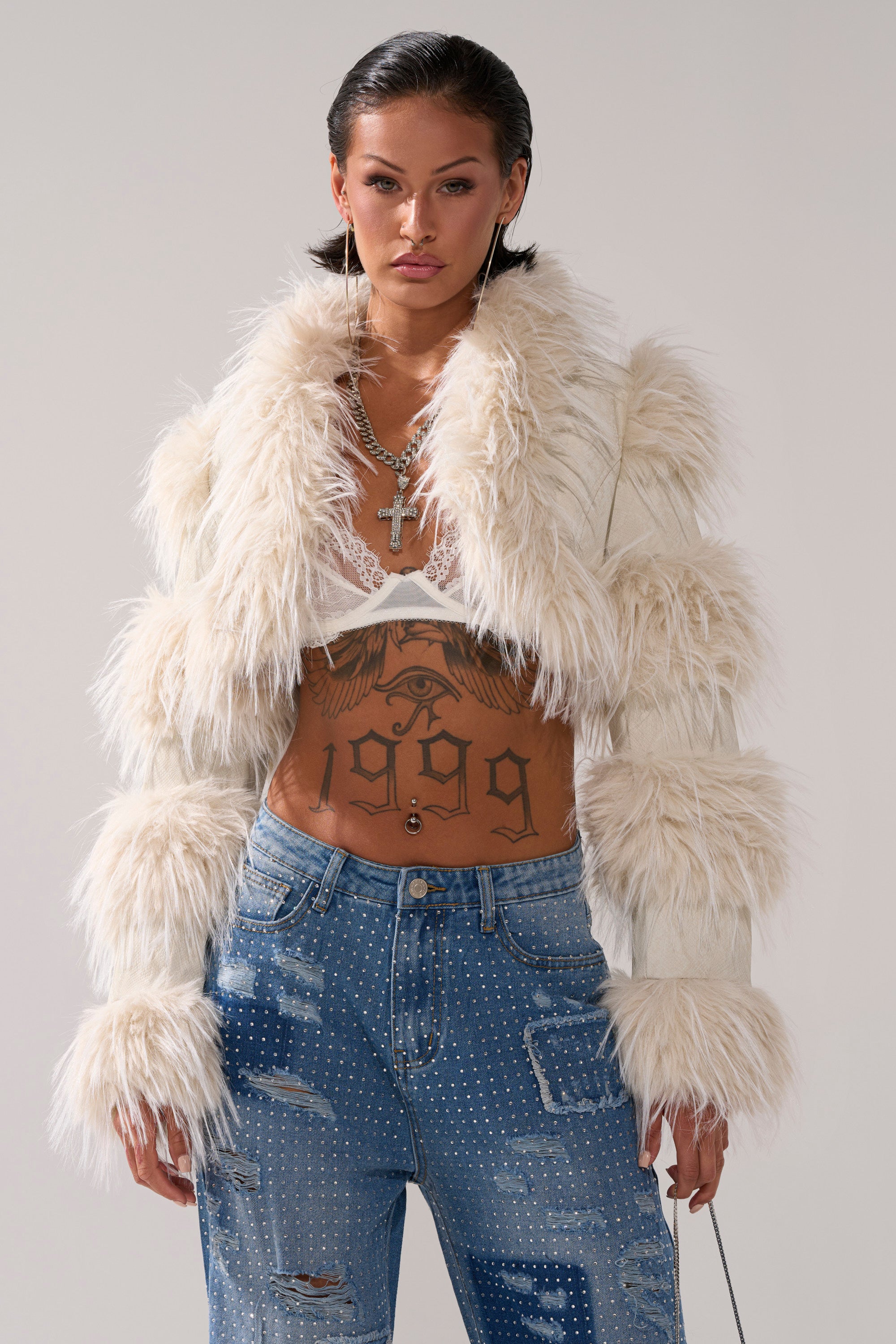 Wearing the LOVERGIRL ULTRA CROP FUR TRIM MOTO IN IVORY over a lace bralette and distressed blue jeans with rhinestones, this person faces the camera neutrally, layered necklaces and a large "1909" torso tattoo on display.