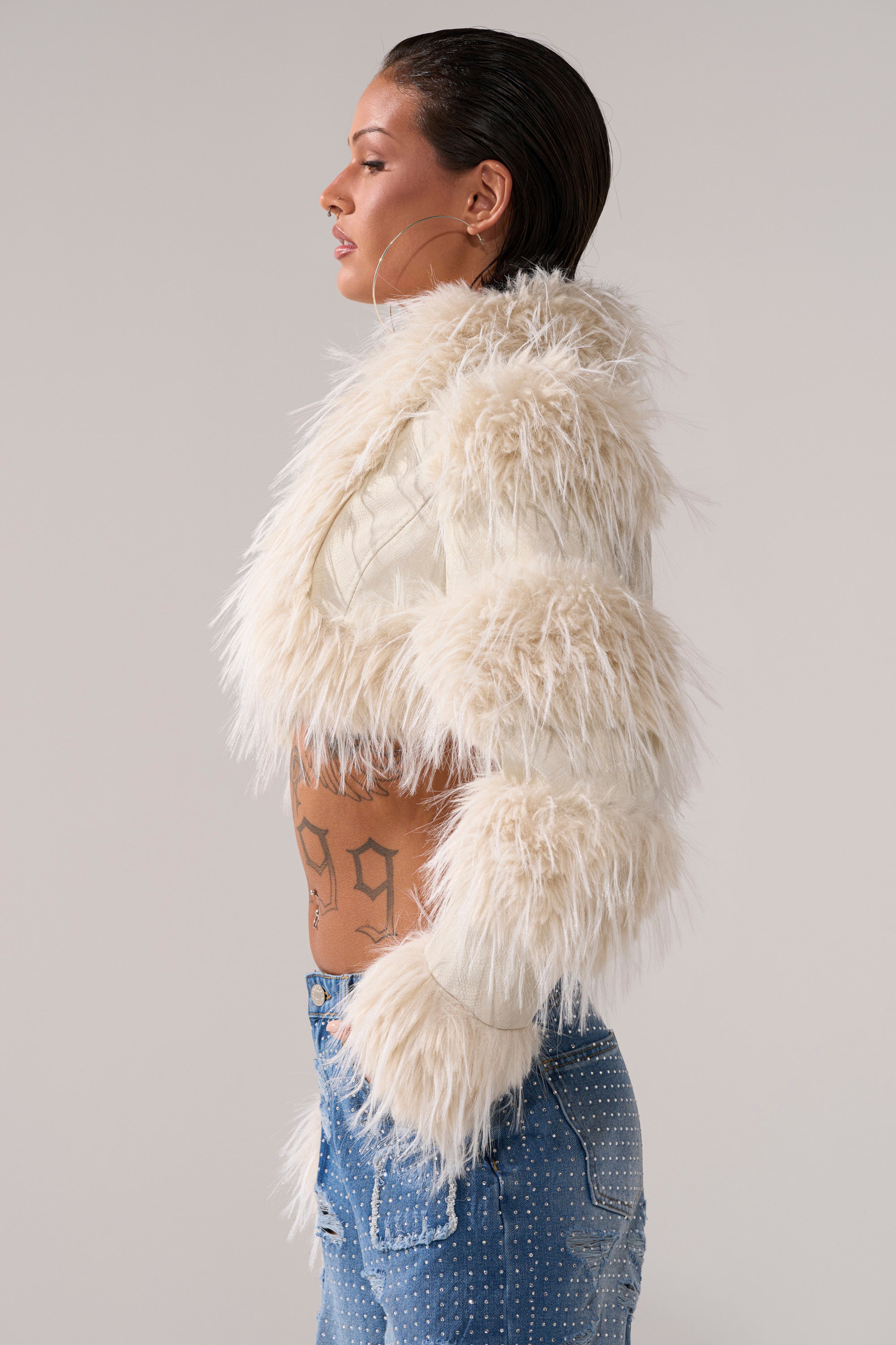 A woman in profile wears the LOVERGIRL ULTRA CROP FUR TRIM MOTO IN IVORY, large hoop earrings, and blue studded jeans. She has slicked-back hair, visible side and lower back tattoos, against a plain light gray background.
