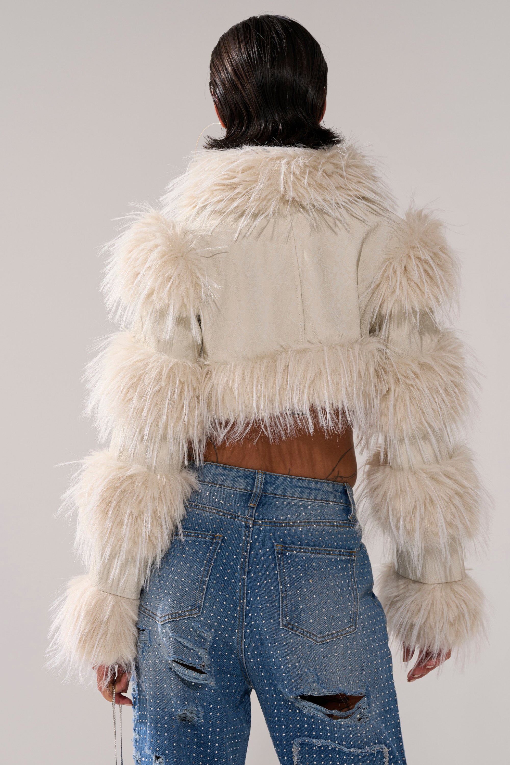 A person with slicked-back dark hair is seen from behind wearing the LOVERGIRL ULTRA CROP FUR TRIM MOTO IN IVORY, paired with light blue ripped jeans adorned with silver studs, against a plain background.