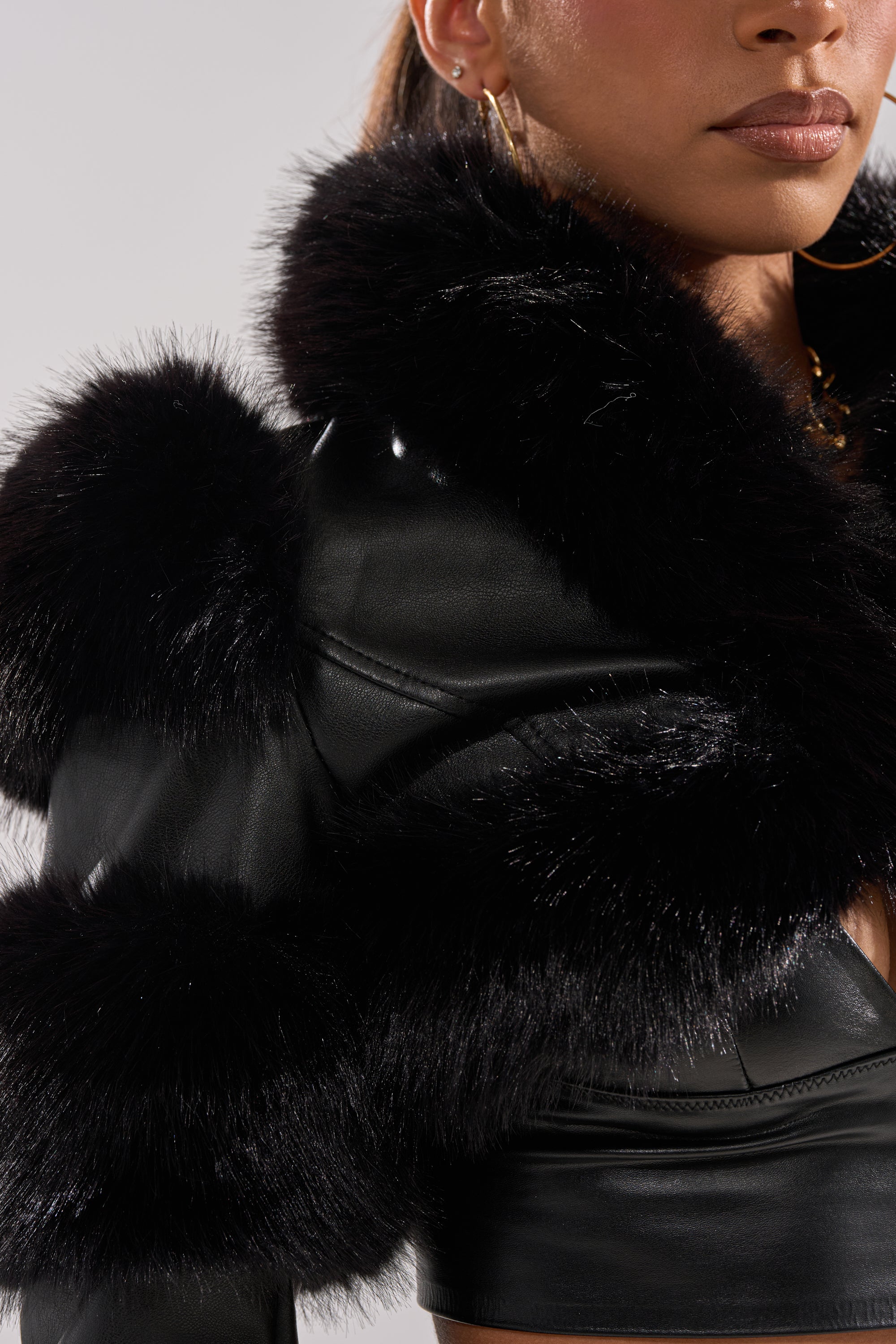 A woman wears the LOVERGIRL ULTRA CROP FUR TRIM MOTO jacket, styled with gold hoop earrings and a gold necklace. The image is cropped to display only the lower half of her face and upper body.