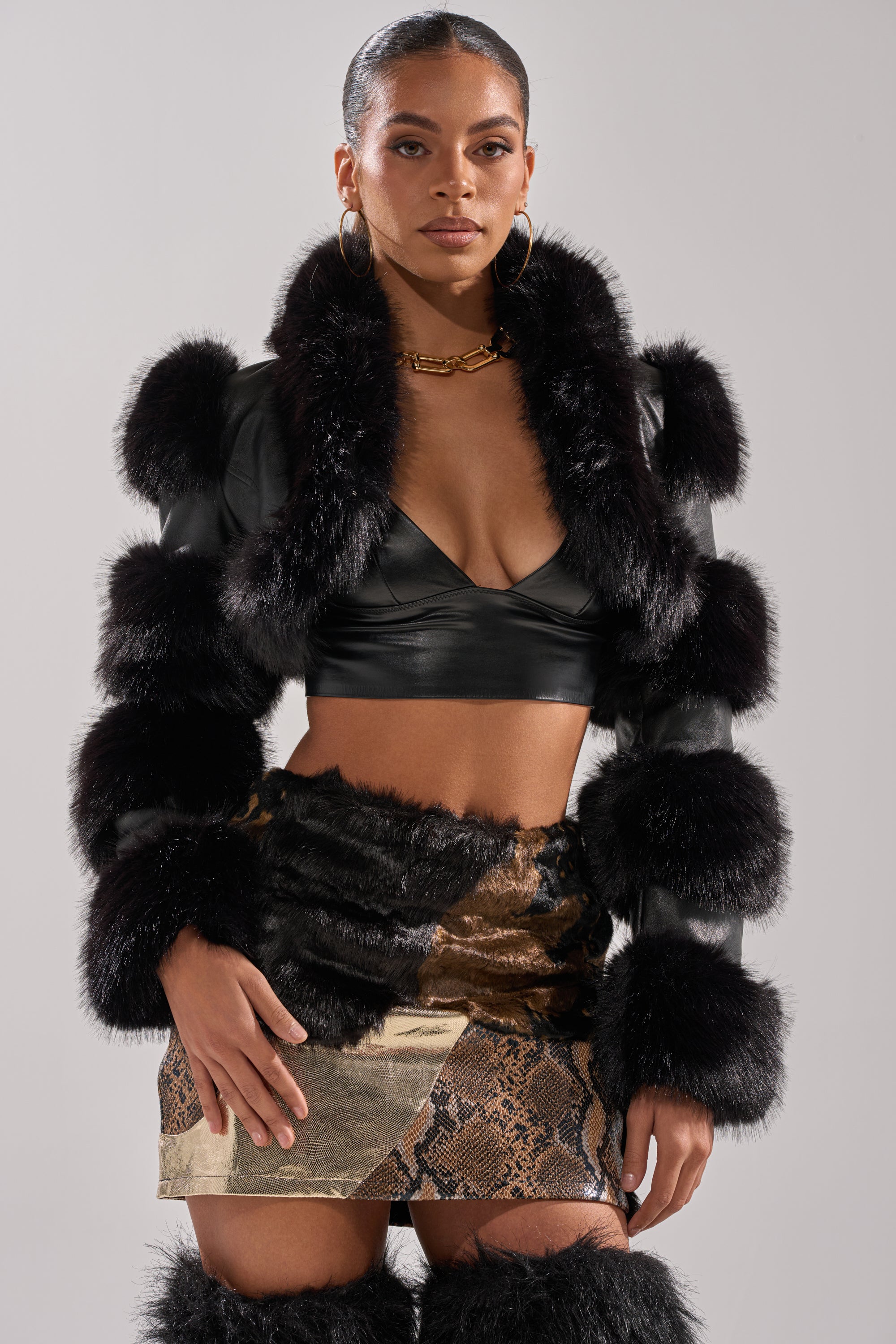 A woman faces forward in the LOVERGIRL ULTRA CROP FUR TRIM MOTO, worn over a black bralette with a metallic snakeskin mini skirt and matching thigh-high boots. Gold hoop earrings and a chunky necklace complete her look.