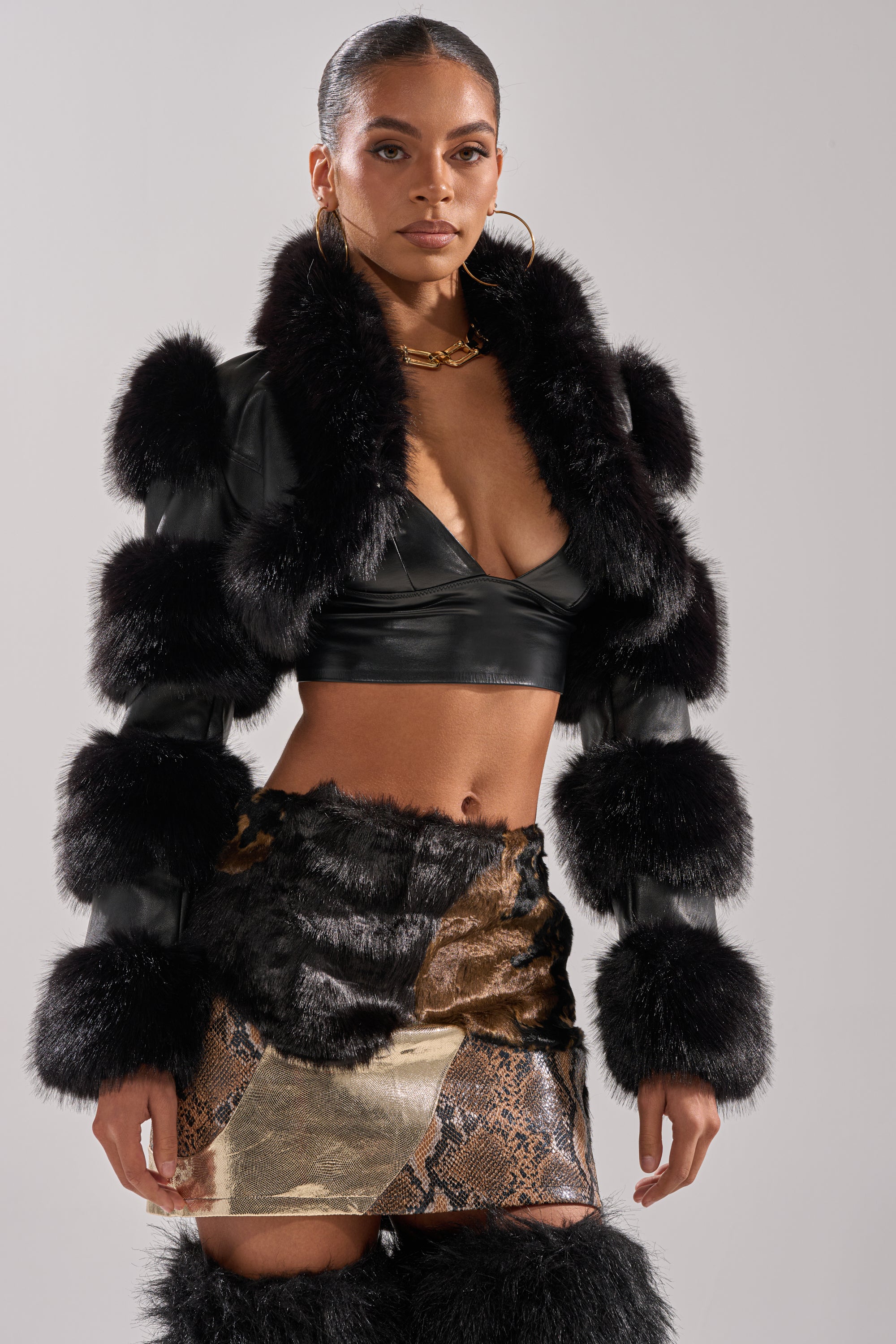 A woman stands confidently against a plain background, styled in the LOVERGIRL ULTRA CROP FUR TRIM MOTO jacket, a black bralette, an animal-print mini skirt with faux fur accents, and large hoop earrings.