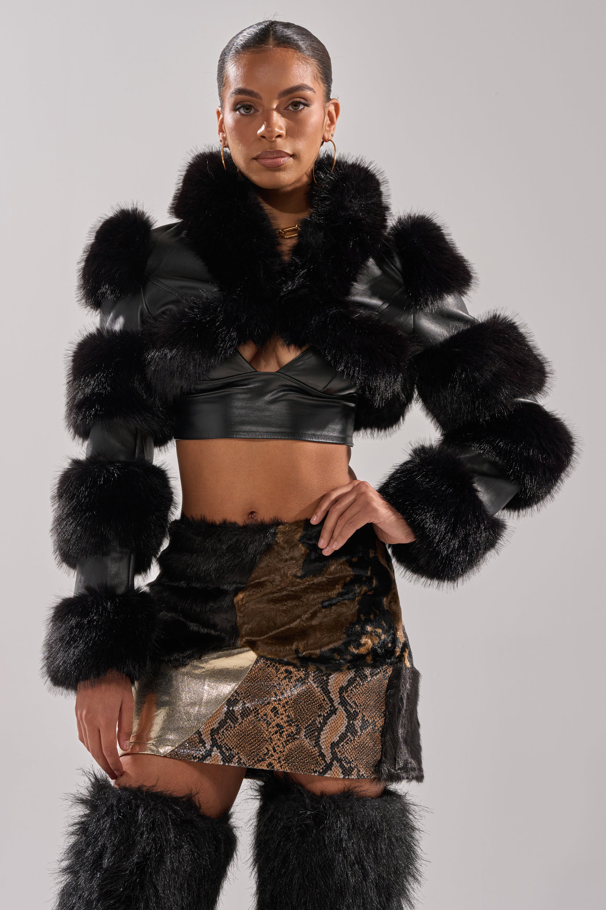 A woman poses confidently in the LOVERGIRL ULTRA CROP FUR TRIM MOTO jacket with black fur, paired with a metallic patchwork snake print mini skirt and matching black fur boots against a light gray background.