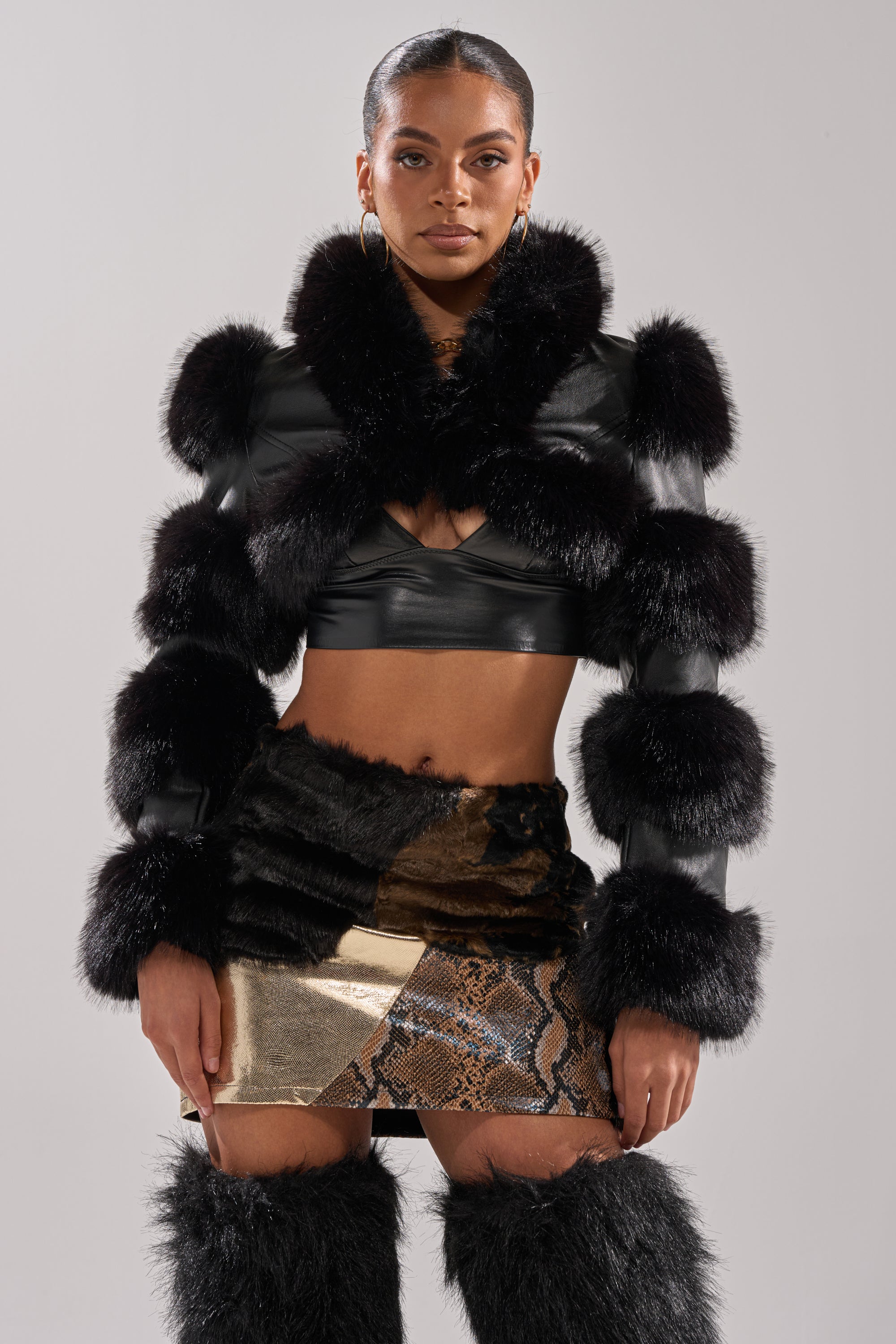 A woman poses confidently in the LOVERGIRL ULTRA CROP FUR TRIM MOTO jacket, paired with a metallic snakeskin mini skirt and matching black furry boots. She wears hoop earrings, has sleek pulled-back hair, and stands against a neutral background.