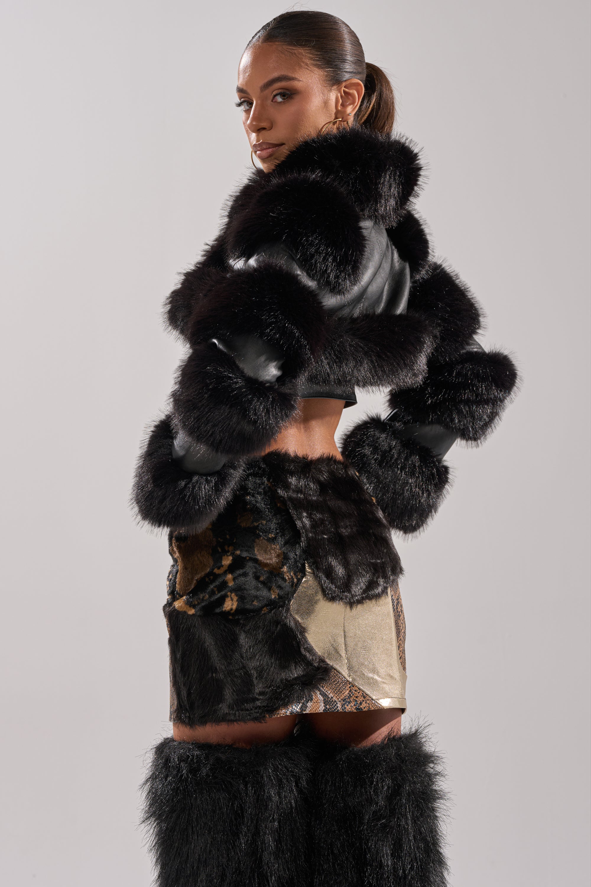 A woman poses in the LOVERGIRL ULTRA CROP FUR TRIM MOTO jacket, pairing it with black fur boots and a textured skirt featuring fur and snake-print patterns, glancing over her shoulder against a plain light backdrop.