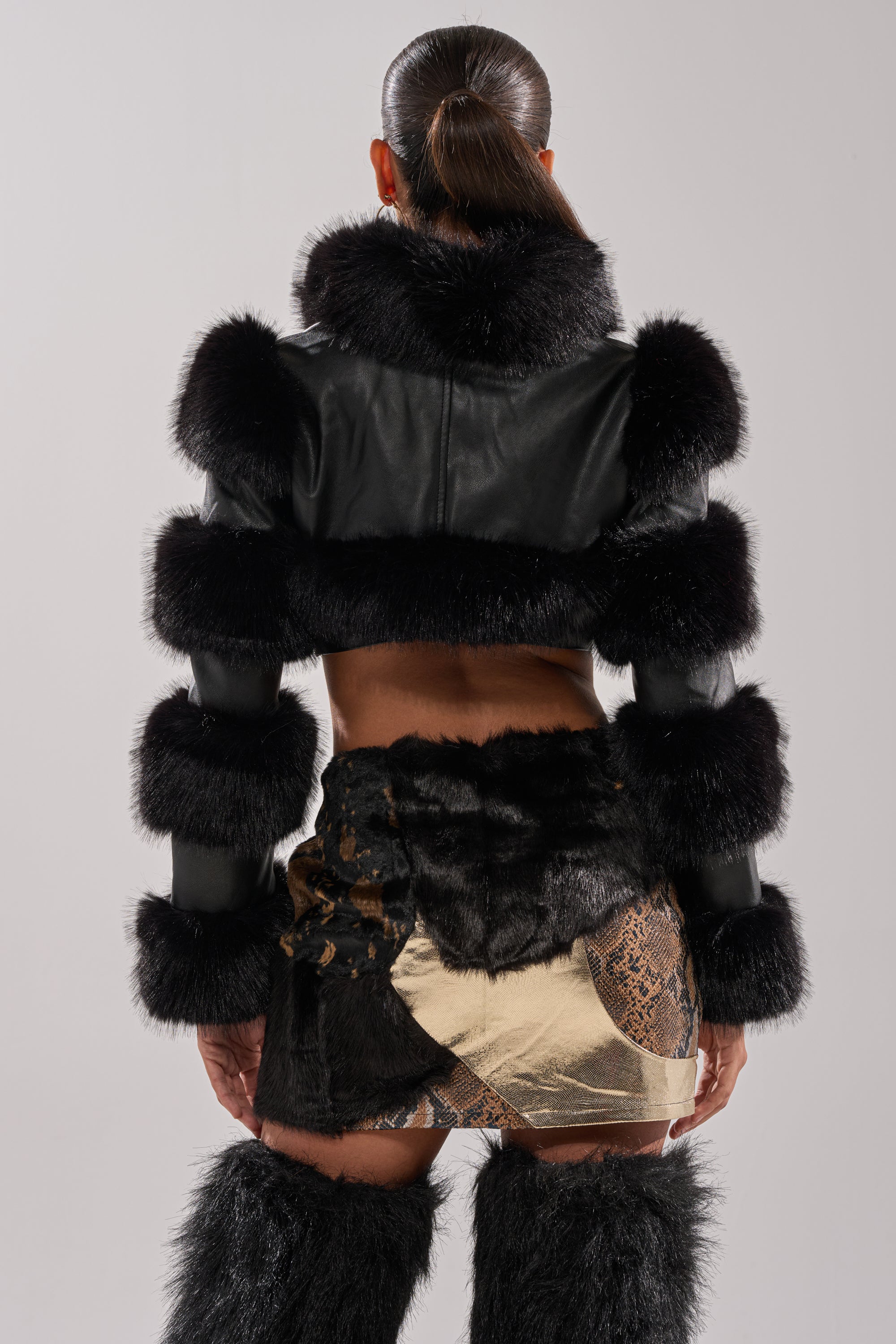 A person faces away in the LOVERGIRL ULTRA CROP FUR TRIM MOTO jacket, a patchwork mini skirt with shiny and textured fabrics, and black fur leg warmers. Their hair is in a sleek ponytail, set against a plain light background.