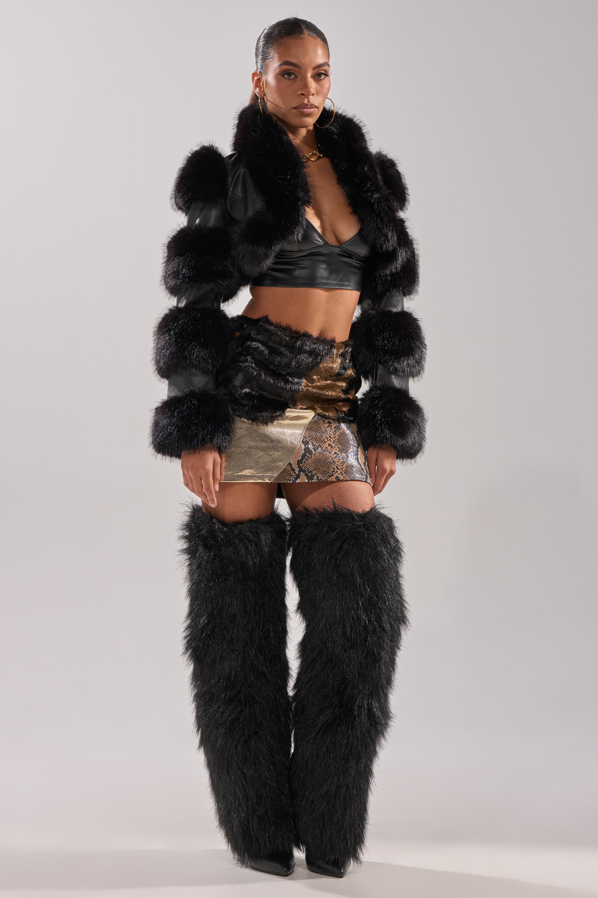 A woman poses in the LOVERGIRL ULTRA CROP FUR TRIM MOTO jacket, styled over a black crop top, gold snakeskin mini skirt, and tall black faux fur boots against a light gray background.