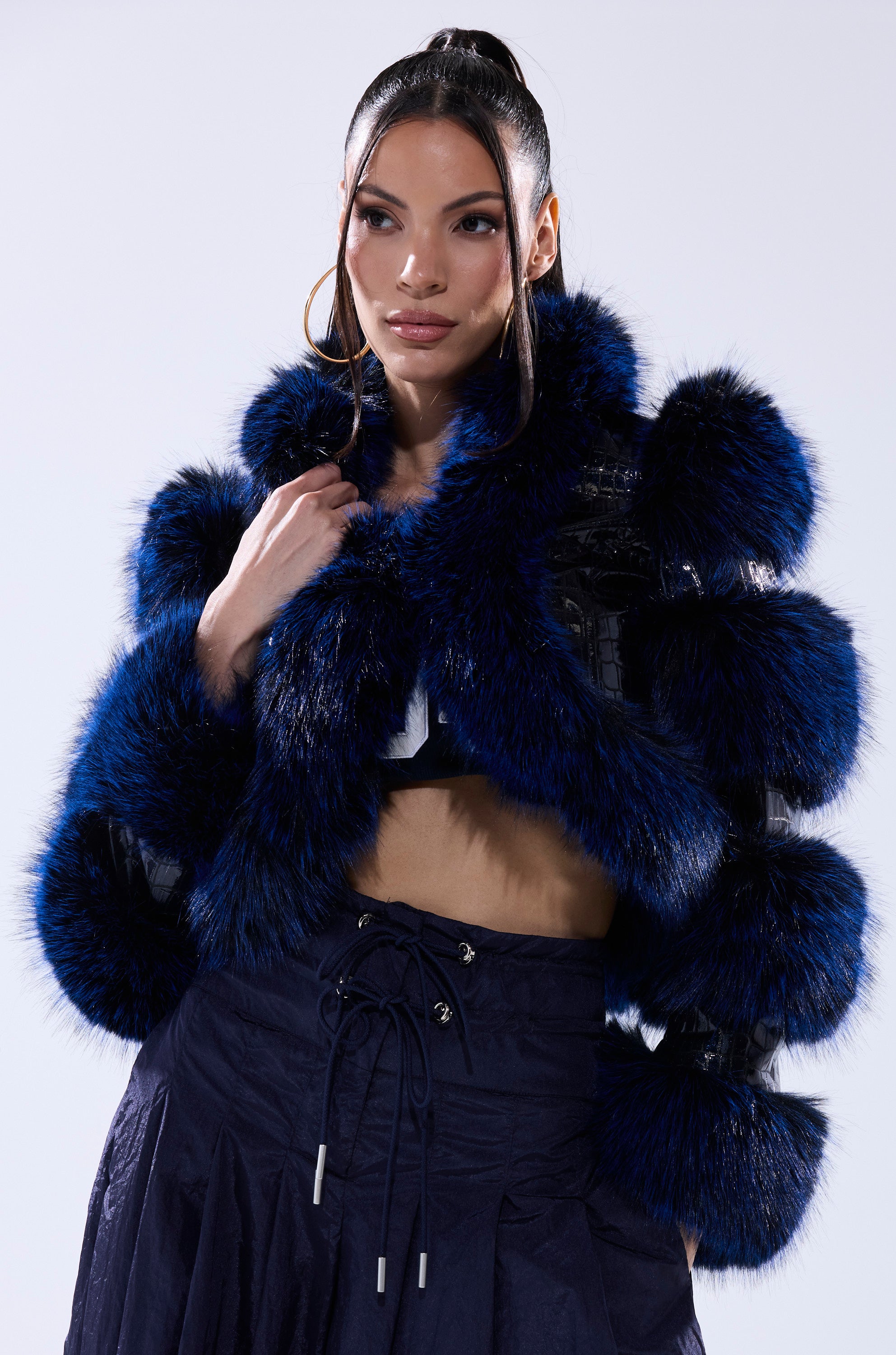 A woman with long dark hair wears the LOVERBOY FUR TRIM MOTO IN NAVY CROC over a black crop top and high-waisted navy pants, standing against a plain, light background.