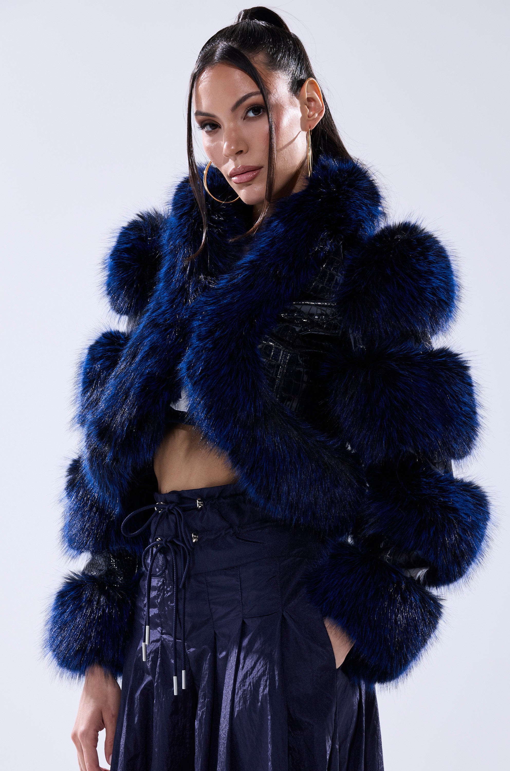 A woman with long dark hair wears the LOVERBOY FUR TRIM MOTO IN NAVY CROC, featuring bold blue and black faux fur accents, paired with a matching navy skirt as she stands confidently against a light background.