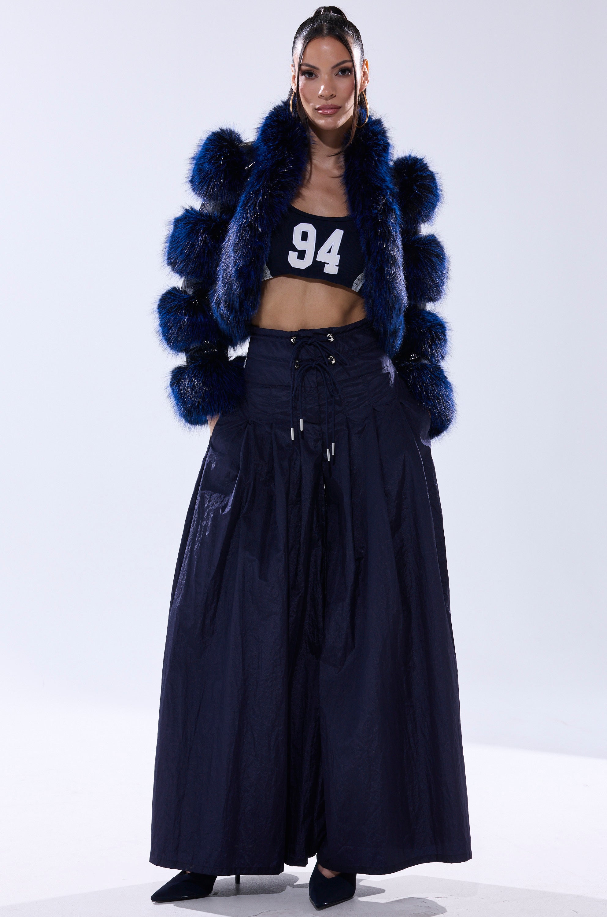 Model stands against a plain background wearing the LOVERBOY FUR TRIM MOTO IN NAVY CROC, a black crop top with "94" in white, high-waisted wide-leg navy pants, and black heels.