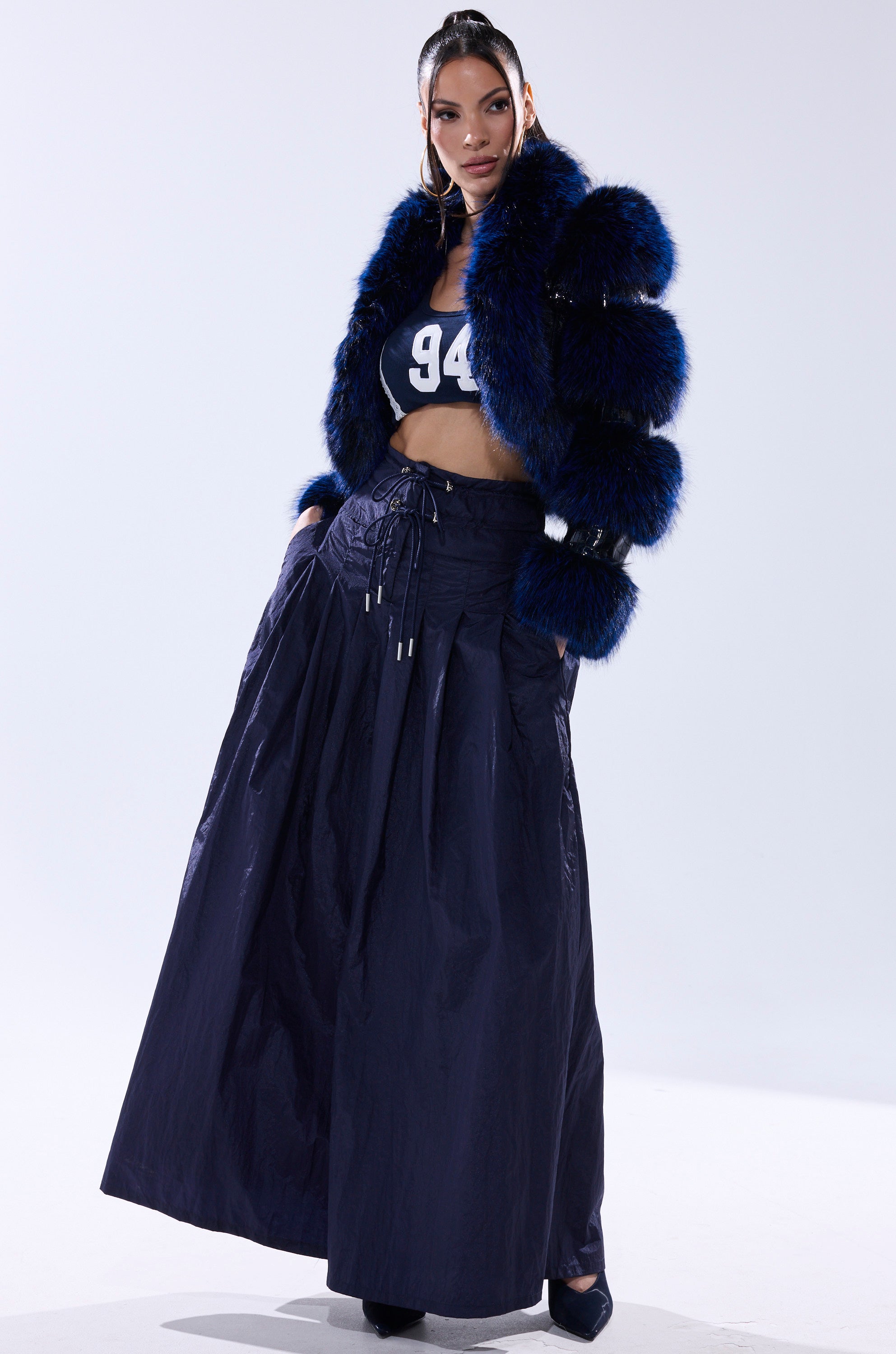 A woman stands confidently in the LOVERBOY FUR TRIM MOTO IN NAVY CROC jacket, paired with a navy sports crop top and long full navy skirt. Her hair is sleek, and she wears dark heels against a plain white background.