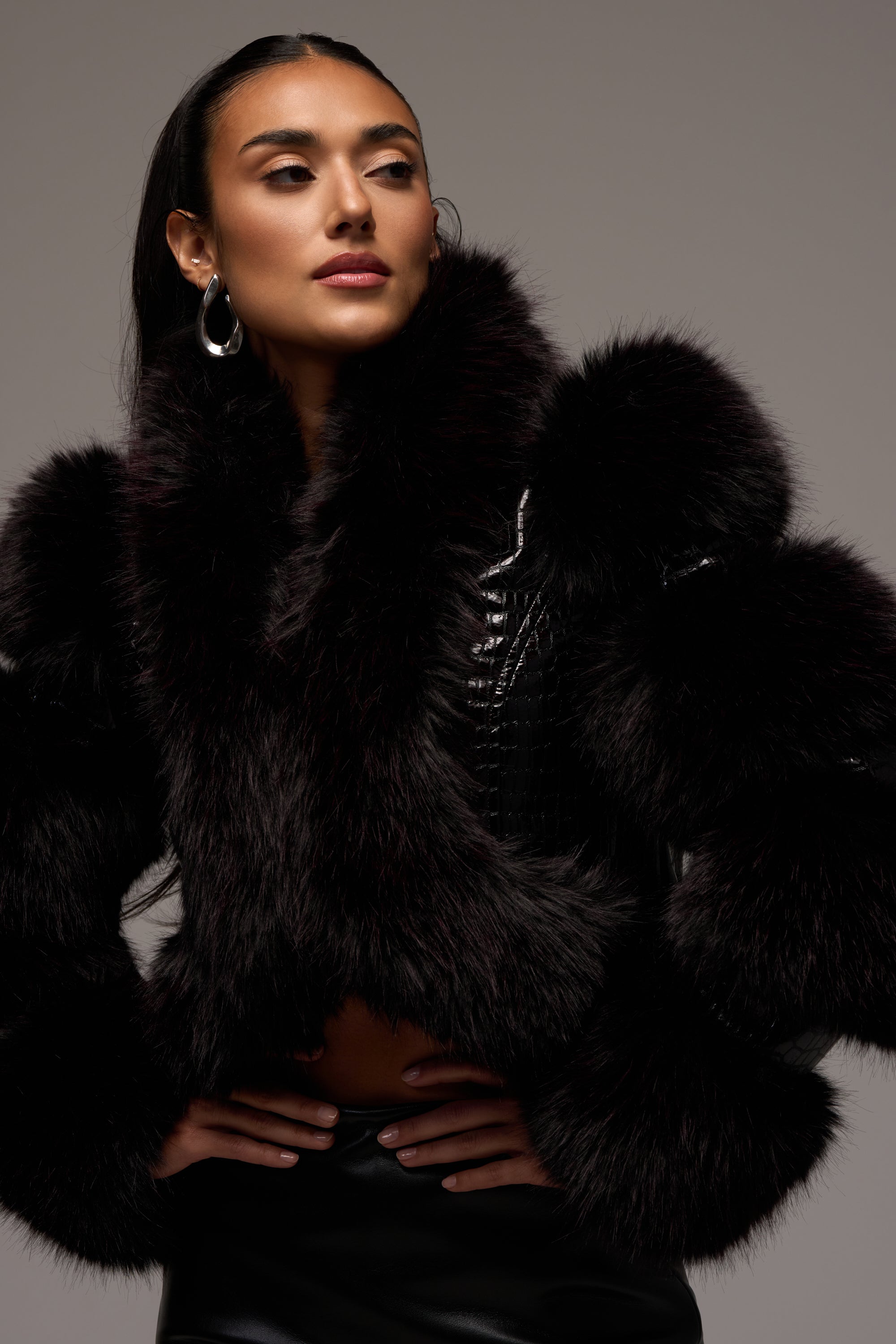 A woman stands against a neutral background, hands on her hips, styled with hoop earrings and a luxurious cropped faux fur jacket featuring voluminous sleeves. She completes the ensemble with the striking LOVERBOY FUR TRIM MOTO IN BURGUNDY CROC—burgundy patent leather boots embossed with a crocodile print.
