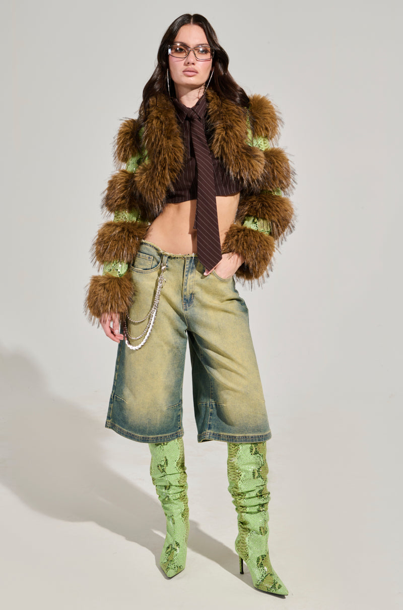 A person poses in the FERNIE ULTRA CROP FUR TRIM MOTO, a furry brown coat with green accents, showcasing its stylish appeal. They complement it with a black crop top and tie, paired with denim knee-length shorts adorned with a decorative chain. Their footwear includes striking green textured knee-high boots featuring a snakeskin pattern. The ensemble is completed with glasses and silver hoop earrings for an added touch of sophistication.
