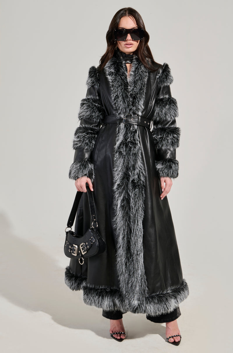 Description: A person wearing the FOR THE DRAMA FUR TRIM COAT featuring a longline silhouette with gray faux fur trim, paired with large sunglasses and black high-heeled sandals. They carry a chic black handbag accented with silver buckles. The backdrop is a simple, light gray.