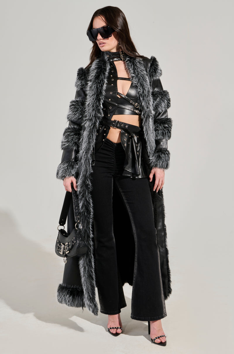 Wearing the FOR THE DRAMA FUR TRIM COAT, a striking faux leather piece highlighted by gray and black accents and a longline silhouette, the individual exudes confidence. They complement the look with stylish sunglasses, elegant heeled sandals, and a small black handbag as they pose against a simple backdrop.