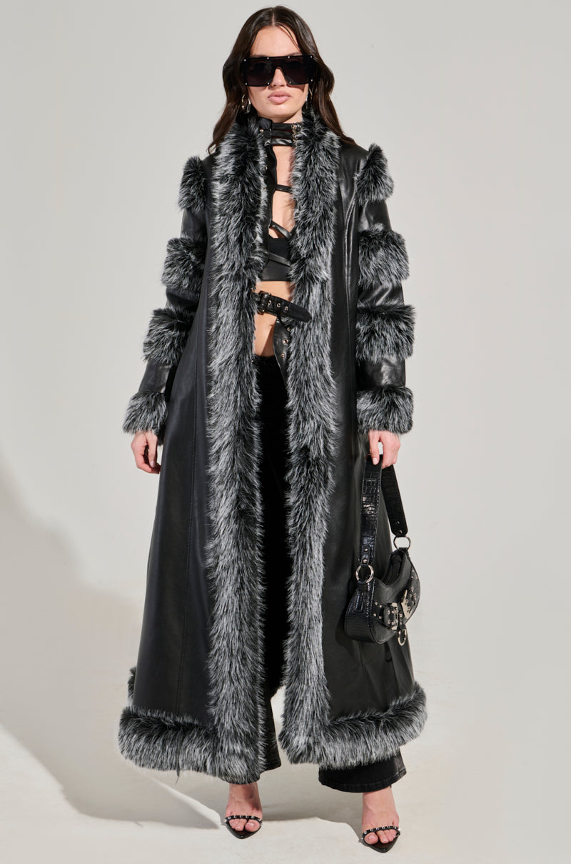 A person confidently stands wearing the "FOR THE DRAMA FUR TRIM COAT," a long black coat featuring chic gray faux fur trim. Their look is complemented by their long dark hair, stylish sunglasses, and a sleek black bag. With the coat open, it reveals a strappy top that enhances the longline silhouette, paired with elegant black pants and heels.