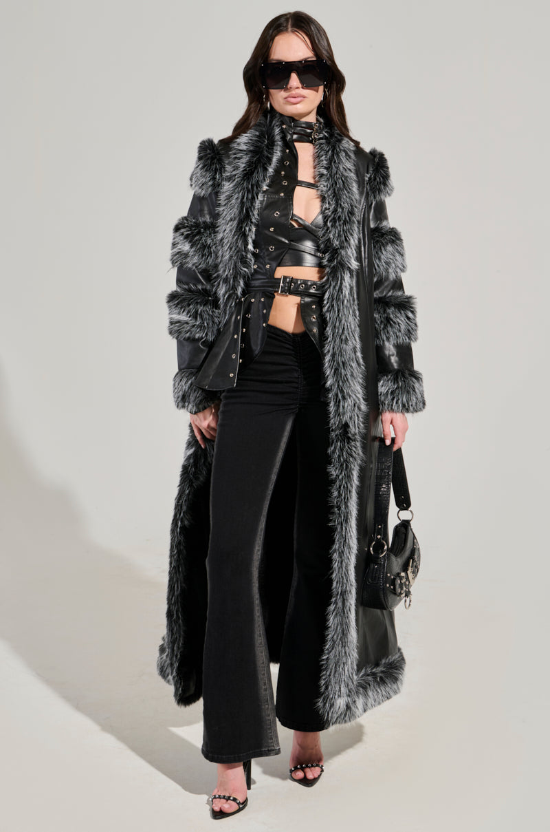 A person wearing the FOR THE DRAMA FUR TRIM COAT, a long black faux leather coat with a chic gray fur trim and sleek longline silhouette, paired with matching black pants and a cropped top. They accessorize with large sunglasses and hold a stylish black handbag against the plain light background.