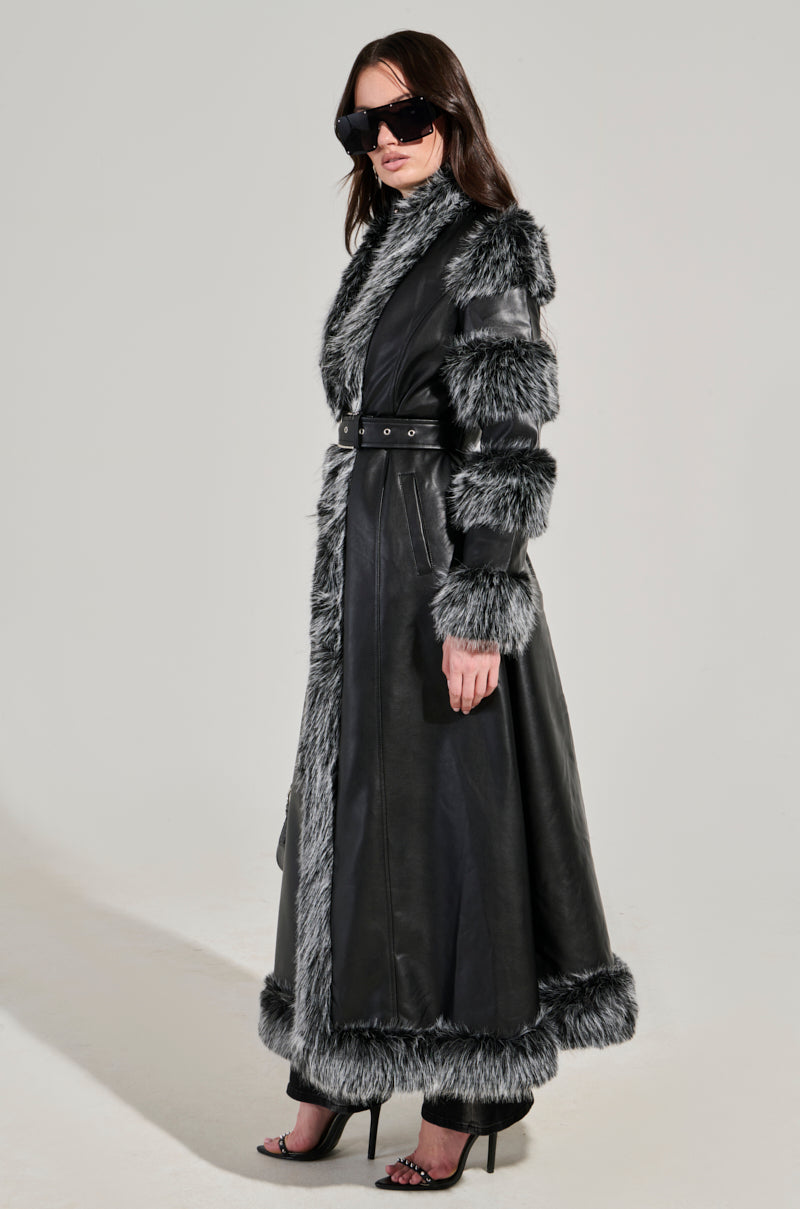 Dressed in the FOR THE DRAMA FUR TRIM COAT, a striking longline silhouette in black faux leather with gray fur trim, the individual exudes confidence. Paired with dark sunglasses and high heels, they pose against a plain backdrop.