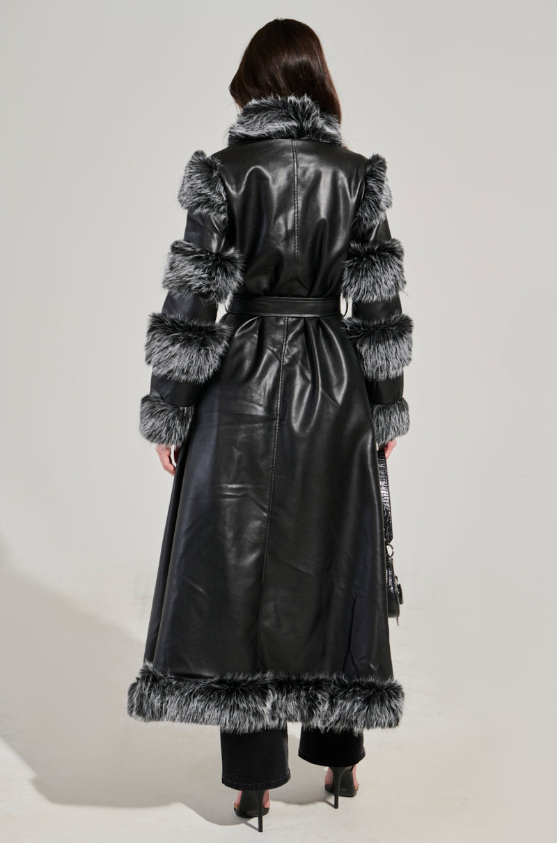 A woman is modeling the FOR THE DRAMA FUR TRIM COAT, featuring a longline silhouette in faux leather with gray fur trim accents on the shoulders, sleeves, and hem. She stands facing away against a neutral background while holding a small black purse.