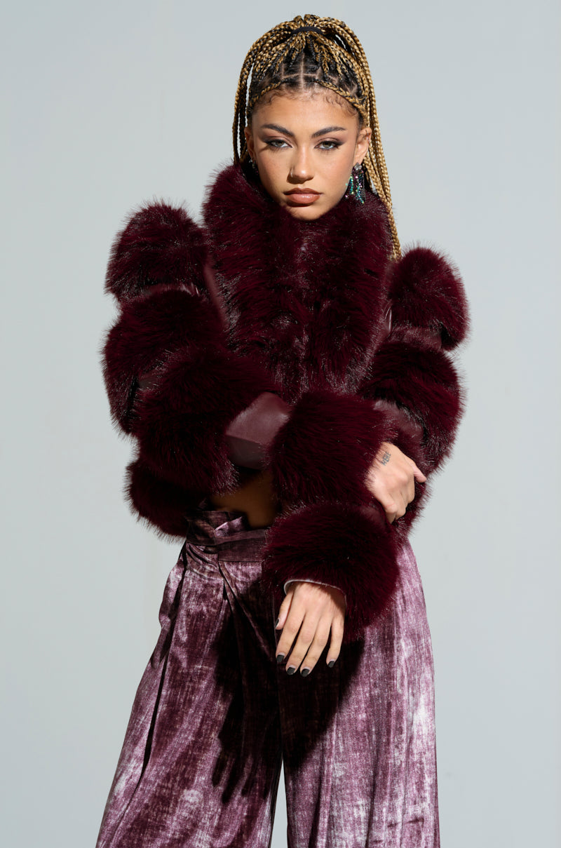 A person with braided hair poses confidently against a plain backdrop, showcasing the stylish LOVERBOY Faux Fur Moto Jacket in burgundy paired with velvet pants. One hand is tucked into the jacket while the other rests on their arm.