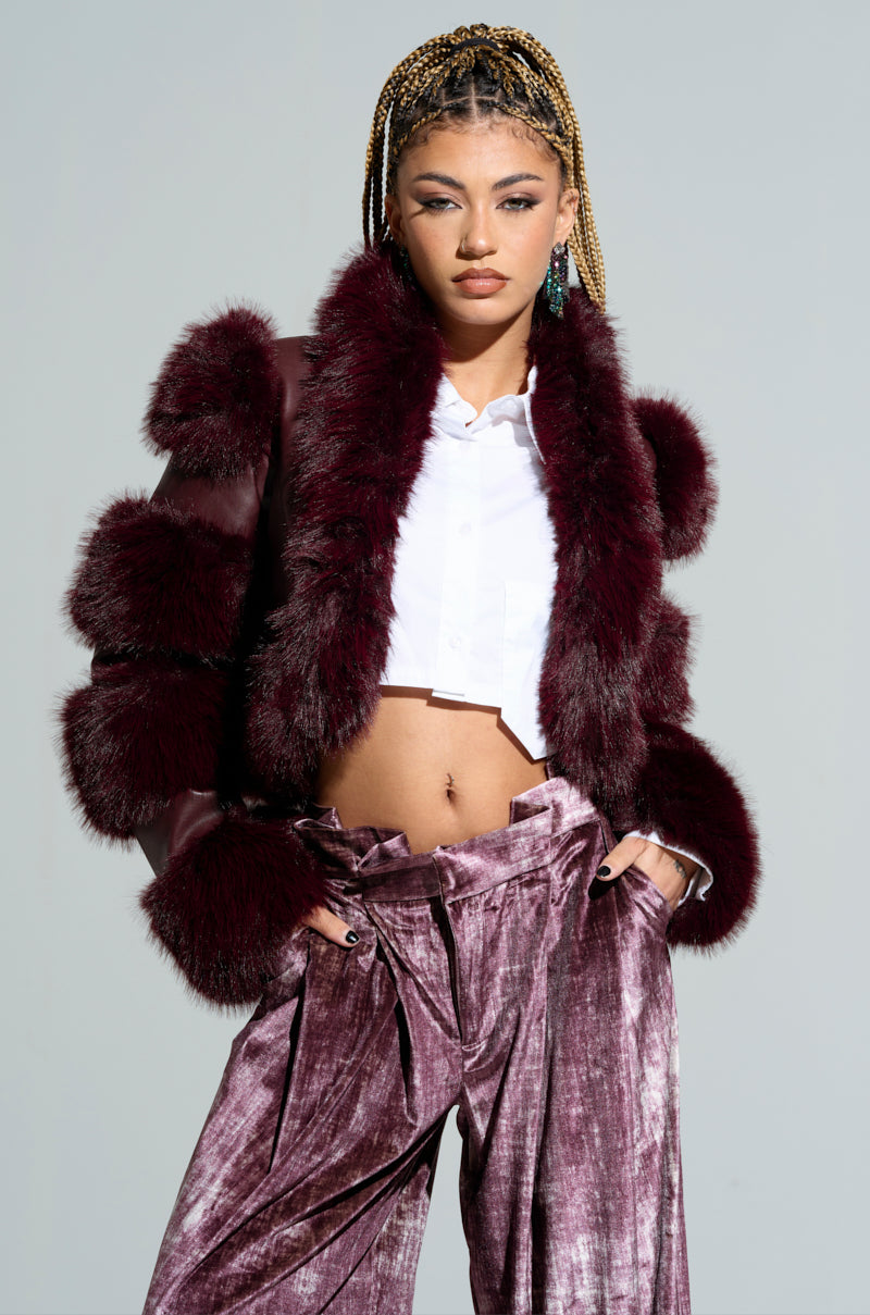 A person with braided hair poses confidently, wearing the Loverboy Faux Fur Moto Jacket in burgundy over a white shirt and high-waisted, velvety pants. Their striking ensemble showcases a rich burgundy hue as they stand against a simple, light gray background.