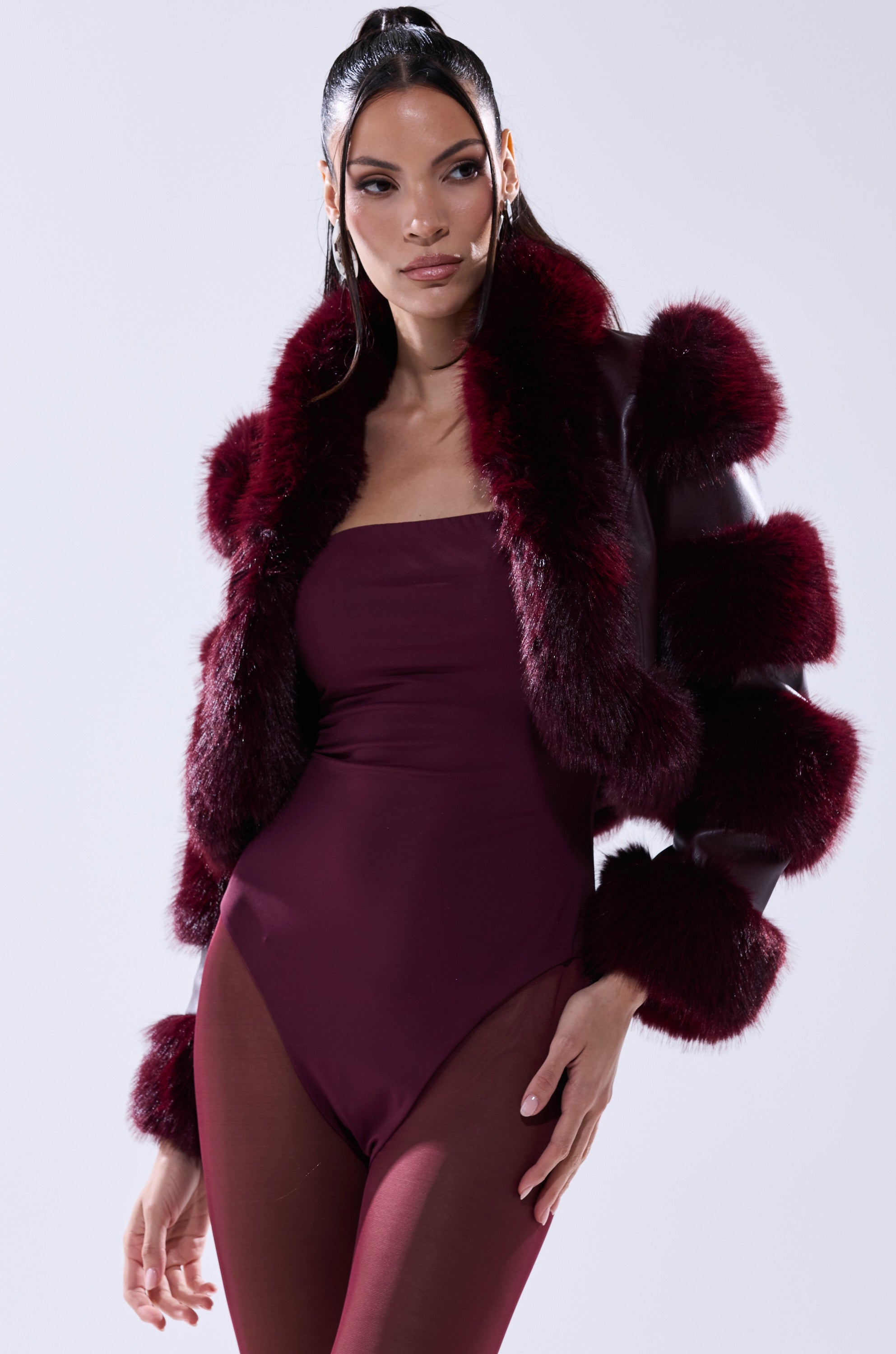 A woman with long dark hair in a high ponytail wears the LOVERBOY FAUX FUR MOTO JACKET IN BURGUNDY, featuring voluminous sleeves, styled over a matching bodysuit against a plain light background.