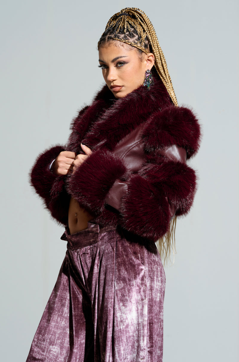 A person with long braided hair poses confidently in the stylish LOVERBOY Faux Fur Moto Jacket in Burgundy, paired with matching velvet pants. Their posture and expression convey a bold and fashionable attitude.